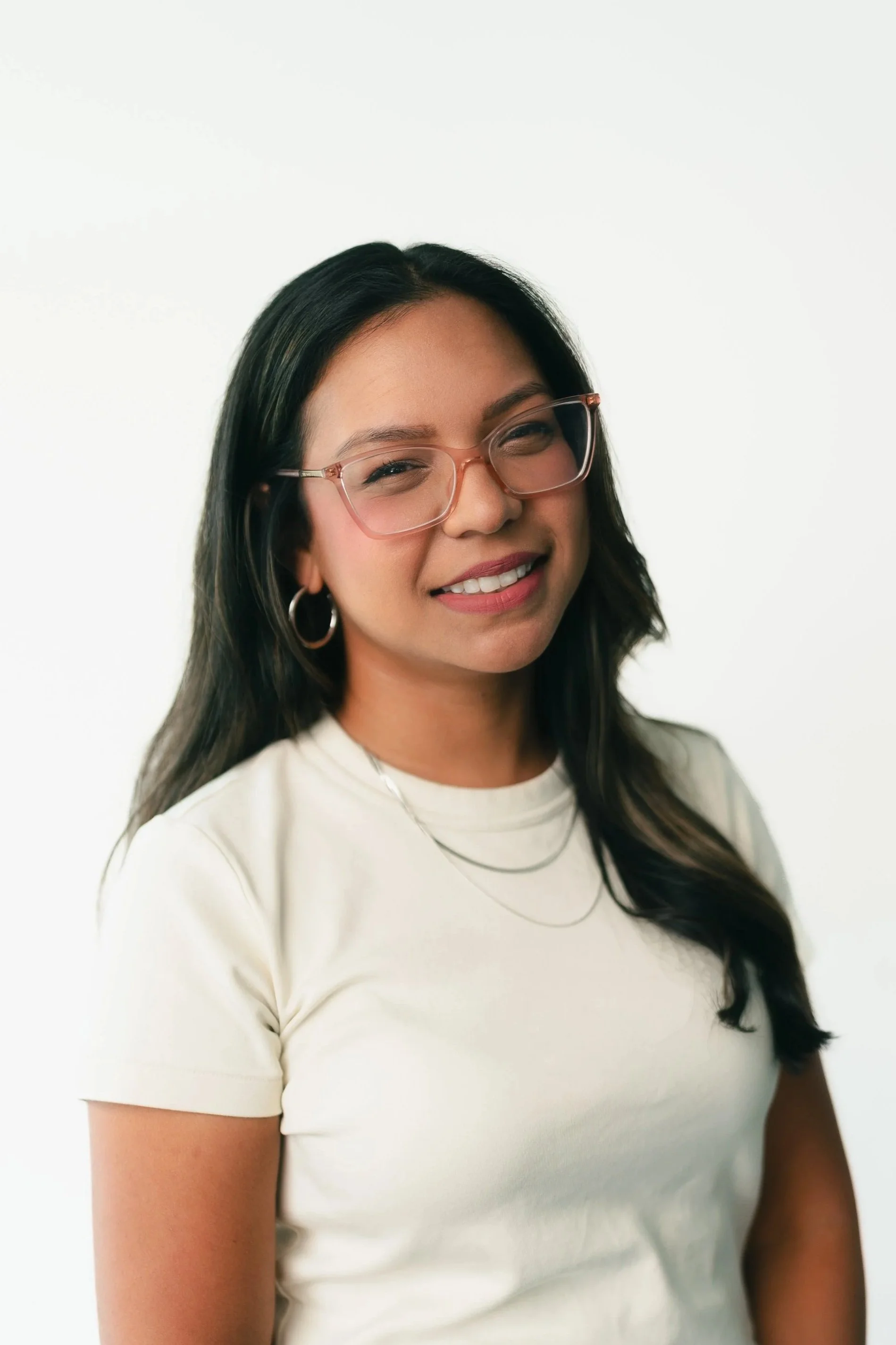Close-up of a young woman with dark hair, wearing glasses, hoop earrings, a white t-shirt, and a delicate necklace, smiling against a plain white background.