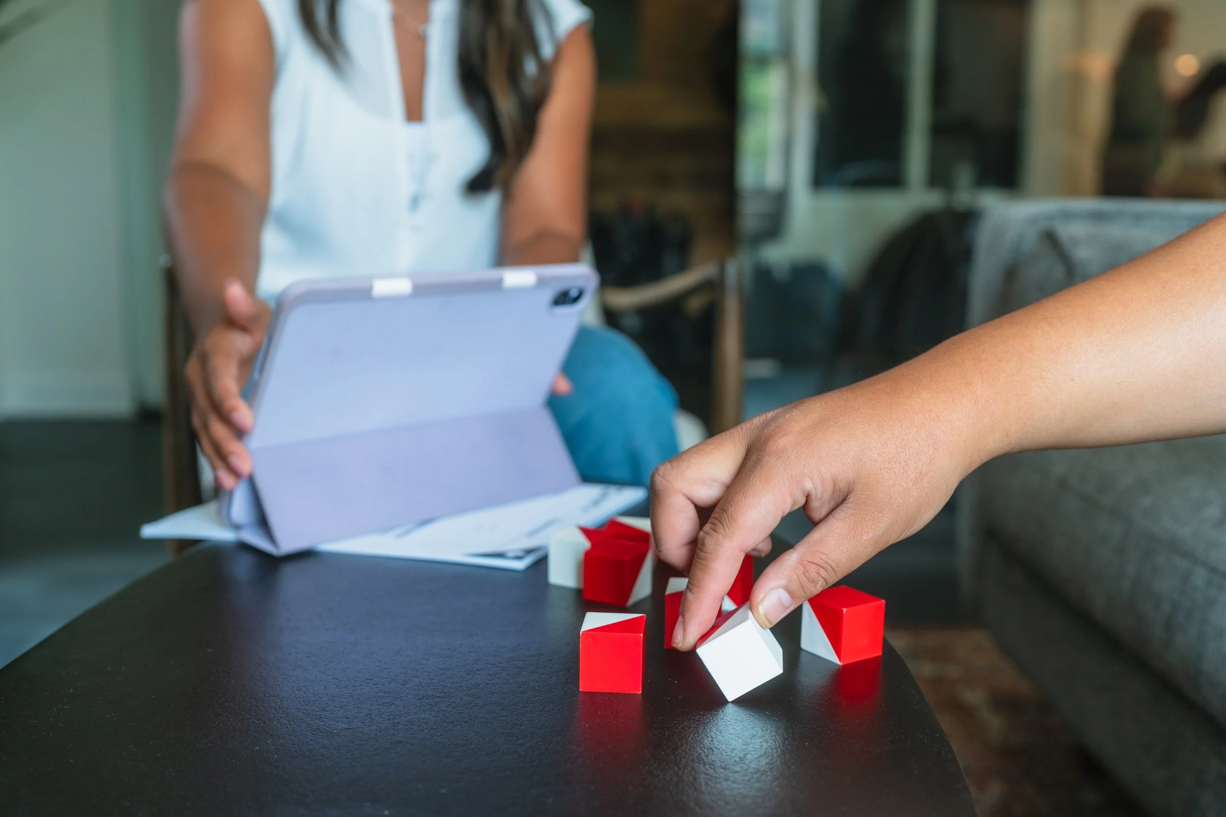 Person playing a tabletop game with red and white blocks on a black table while someone holds a tablet nearby.