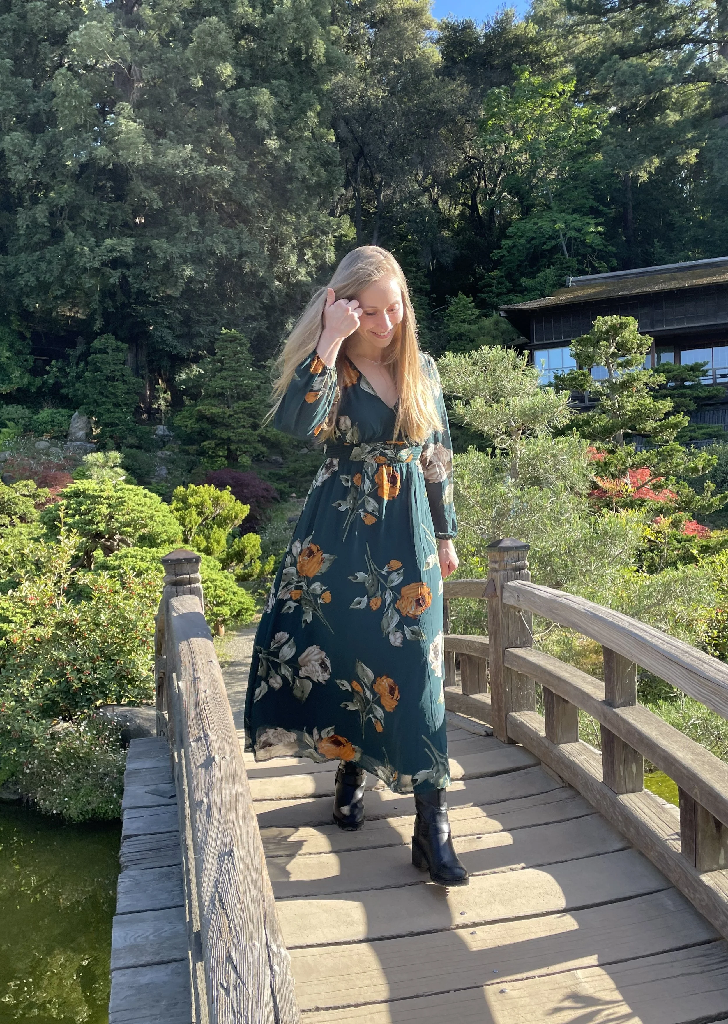 A woman with long blonde hair, wearing a dark teal floral dress and black boots, is walking on a small wooden bridge in a lush, green Japanese-style garden.