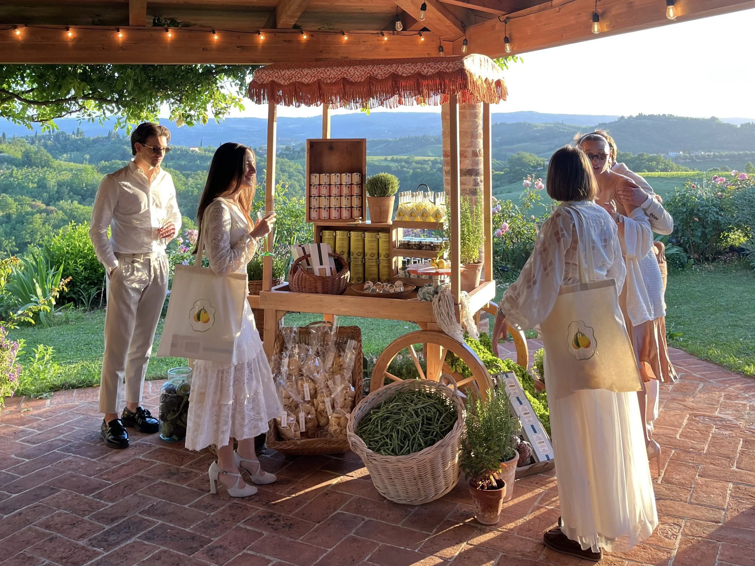 People at an outdoor market stall with a scenic view of hills and trees in the background. The stall has jars, small potted plants, and hanging decorations. The people are dressed in light, elegant clothing and are engaging with each other.