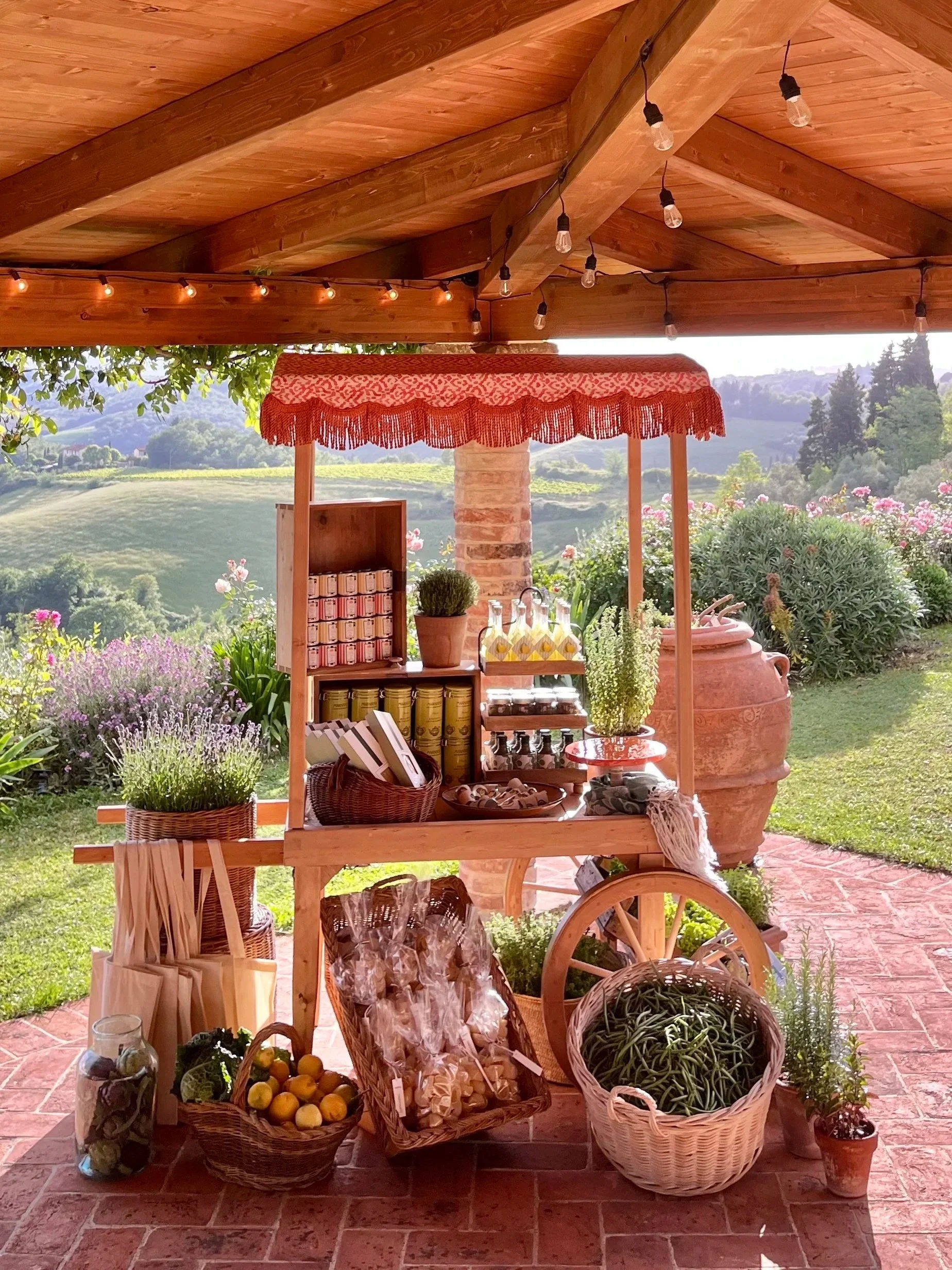 A rustic outdoor market stand with various baskets of fresh produce and jars of preserves, set against a scenic landscape of rolling hills and greenery under a wooden roof adorned with string lights.