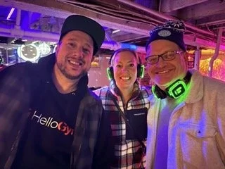 Three smiling people at a social event with colorful lighting, two men and one woman, standing close together.