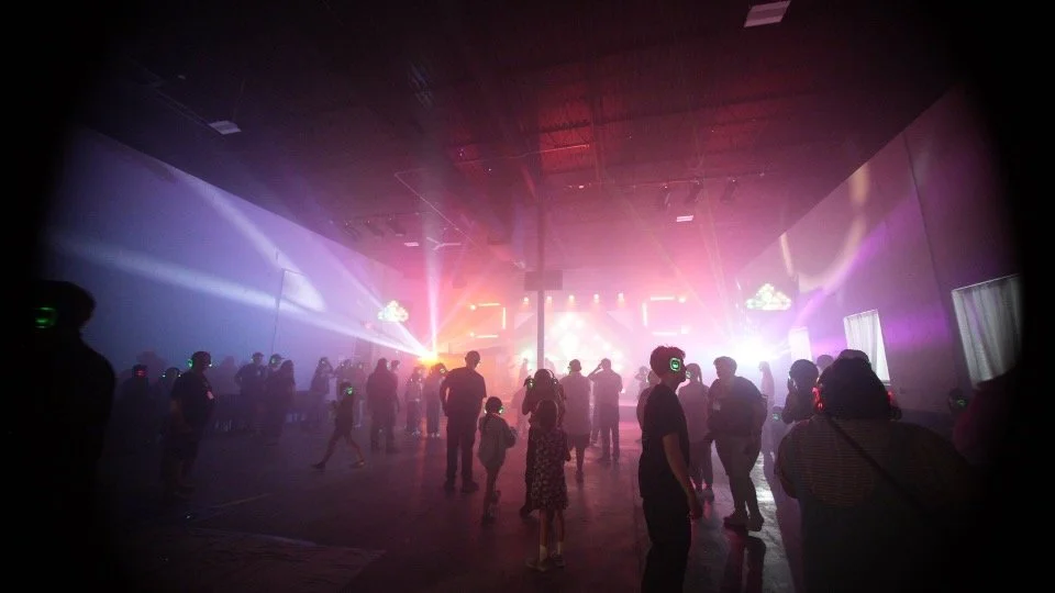 People dancing at a concert or dance event with colorful lights and fog effects.