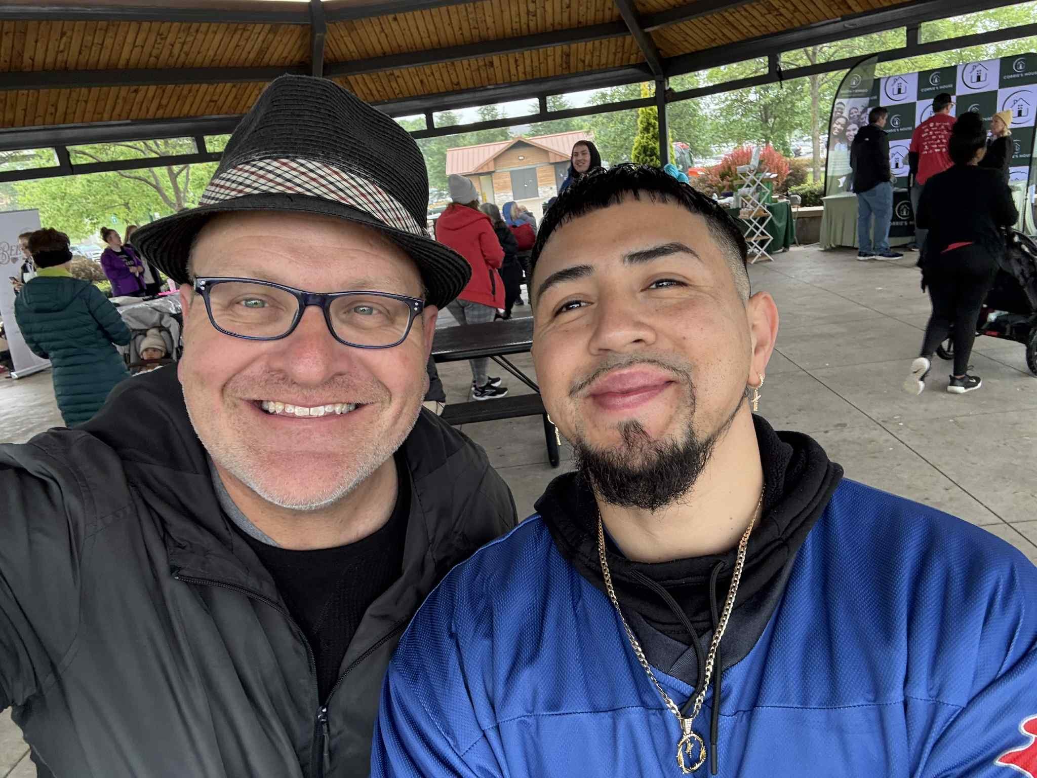 Two men smiling for a selfie at an outdoor event with a pavilion roof. One man has glasses, a black jacket, and a hat. The other has a beard, earrings, and is wearing a blue sports jacket.