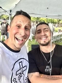 Two men smiling and taking a selfie outdoors under a canopy