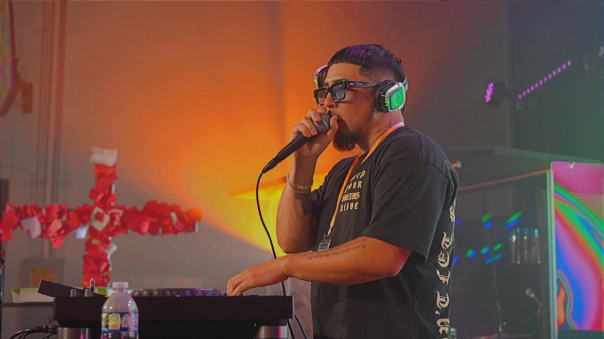 A male DJ wearing sunglasses and headphones is holding a microphone in one hand and operating DJ equipment with the other, standing in front of colorful lighting.