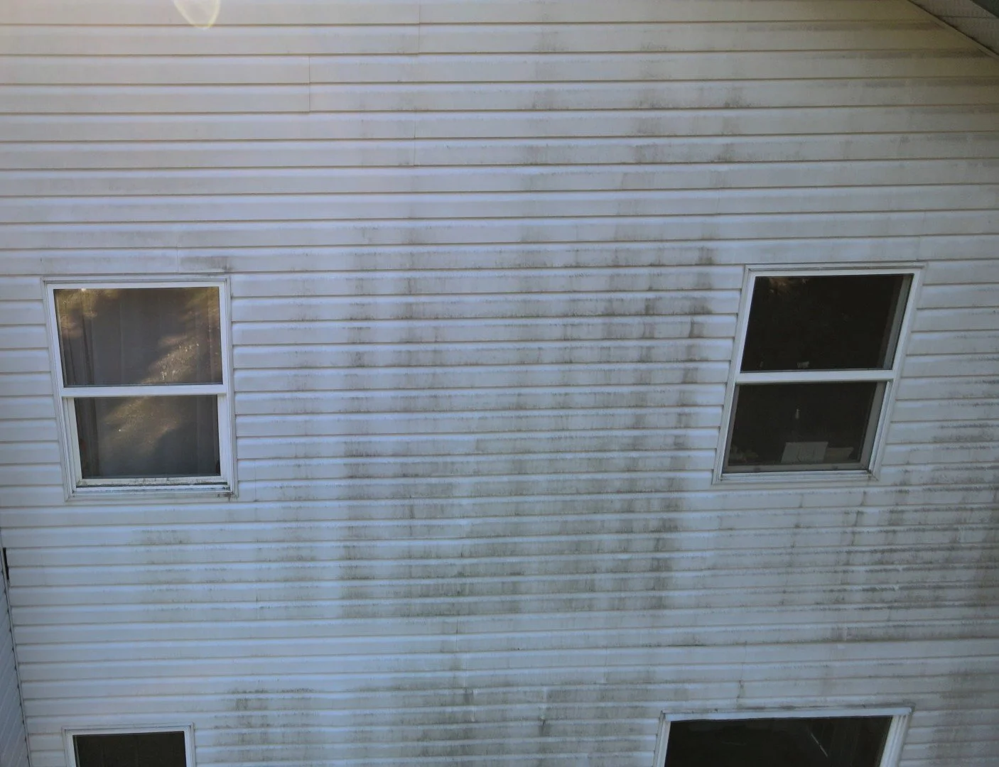The exterior wall of a house with white siding, featuring three windows and visible dirt and grime.