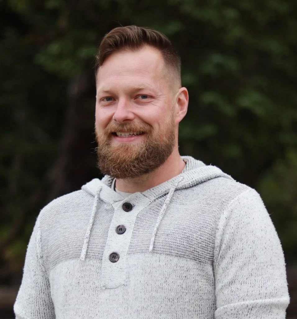 A man with a beard and short hair smiling outdoors, wearing a light-colored hoodie.