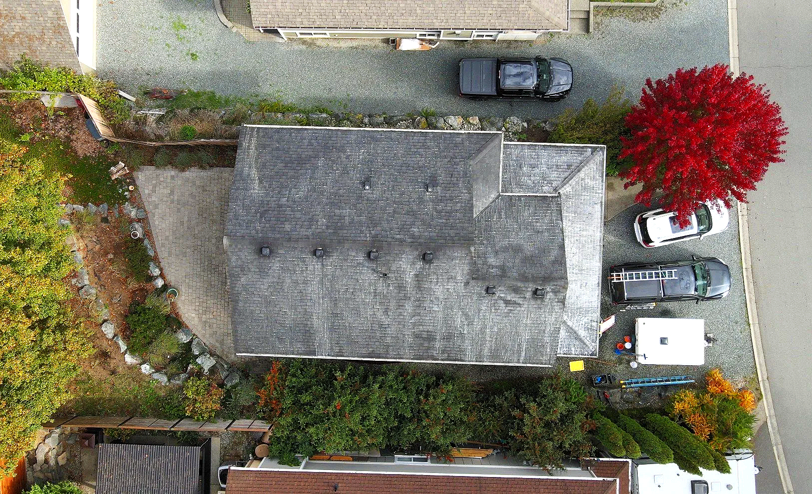 From an aerial view of a residential area showing a house with a gray roof, a driveway with parked cars, a small garden with trees and plants, and neighboring houses and streets.