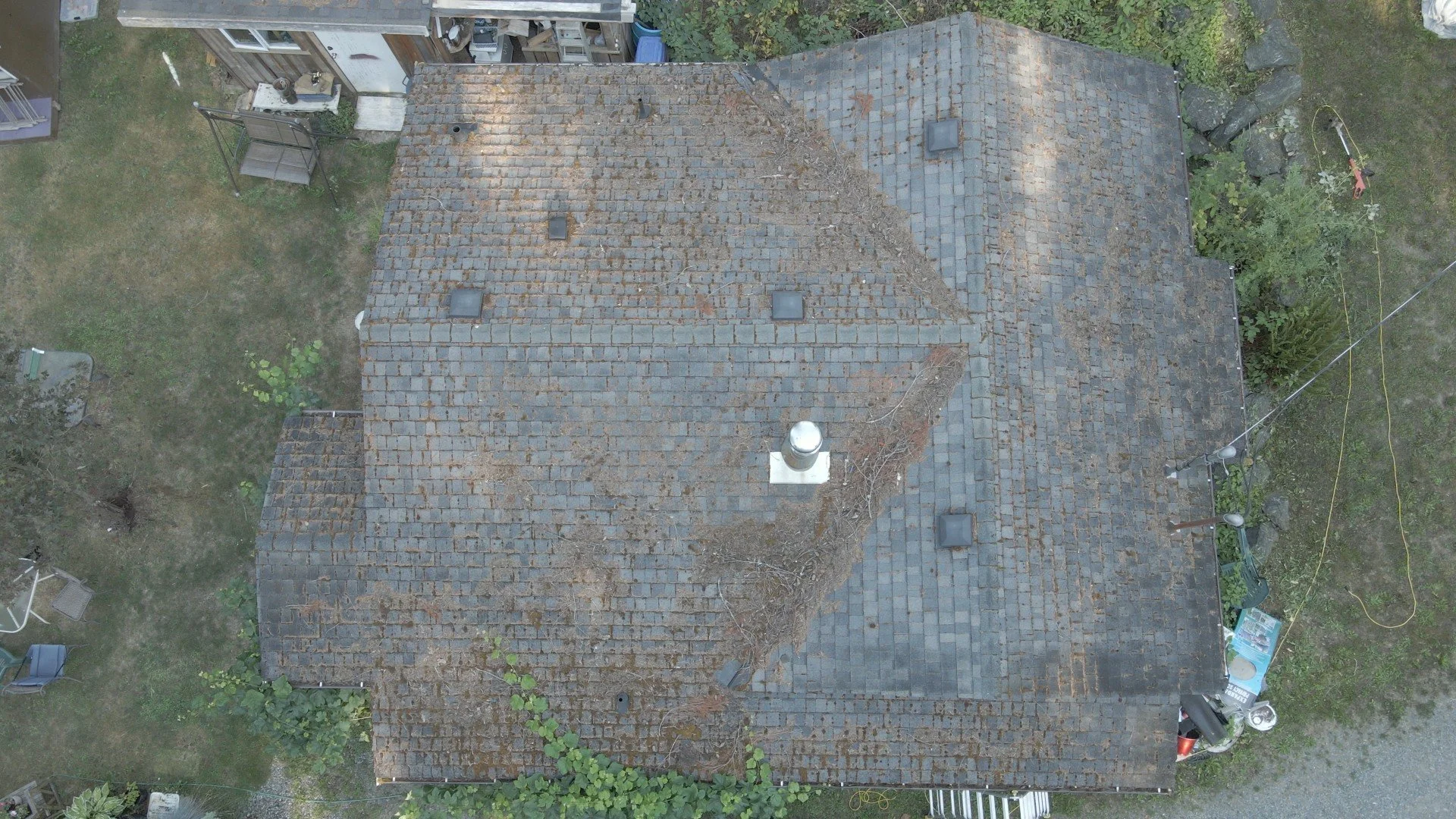 Aerial view of a house roof with shingles, some covered in leaves and debris, and a chimney. Surrounding yard with outdoor furniture, garden, and equipment.