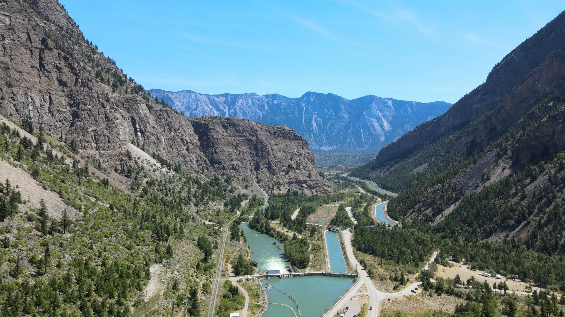 A river with multiple spillways flowing through a mountainous landscape with steep rocky cliffs and forested slopes under a clear blue sky.