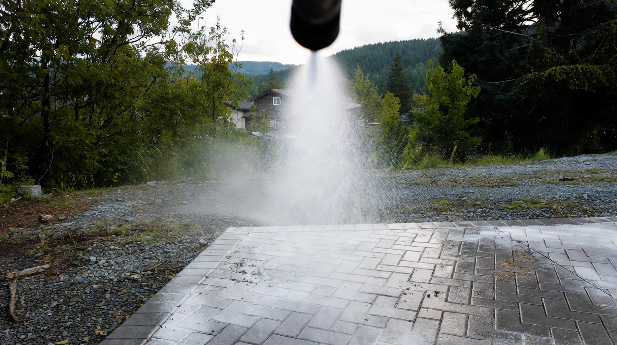 A sprinkler watering gray brick patio, surrounded by green trees and hills in the background.
