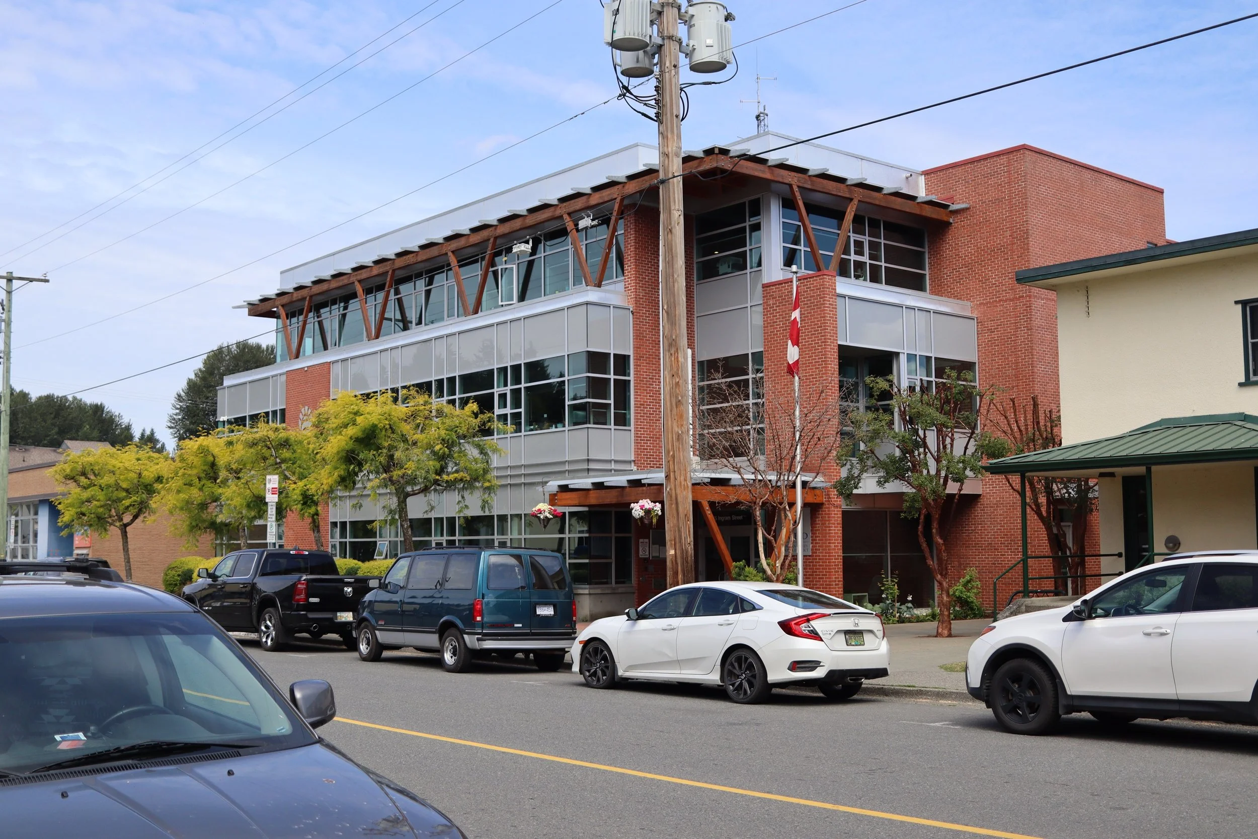 Modern multi-story building with glass windows and brick walls, surrounded by parked cars and trees, under a blue sky.
