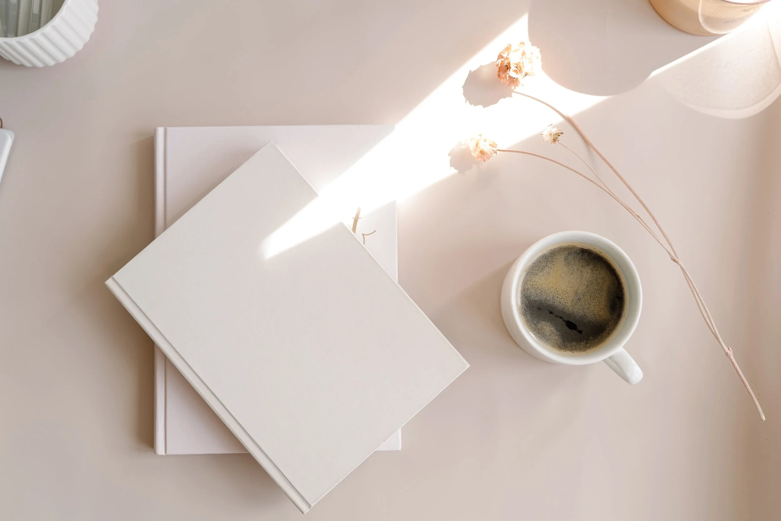 A stack of white closed books, a cup of black coffee, and dried pink flowers on a light-colored table, with sunlight casting a shadow.