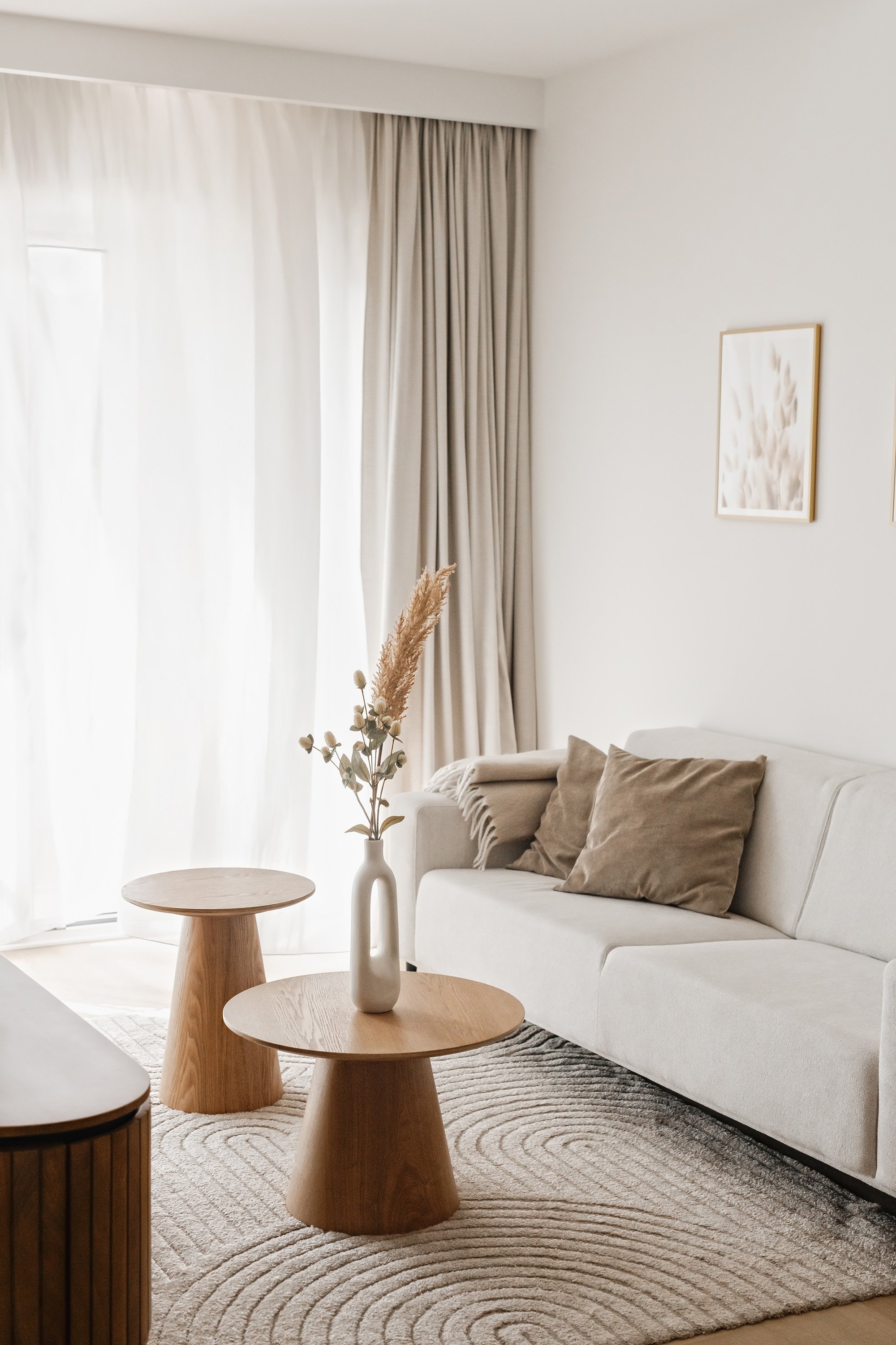 A minimalist living room with beige sofa, matching cushions, wooden side tables, a white vase with dried flowers, sheer curtains, and framed art on a white wall.