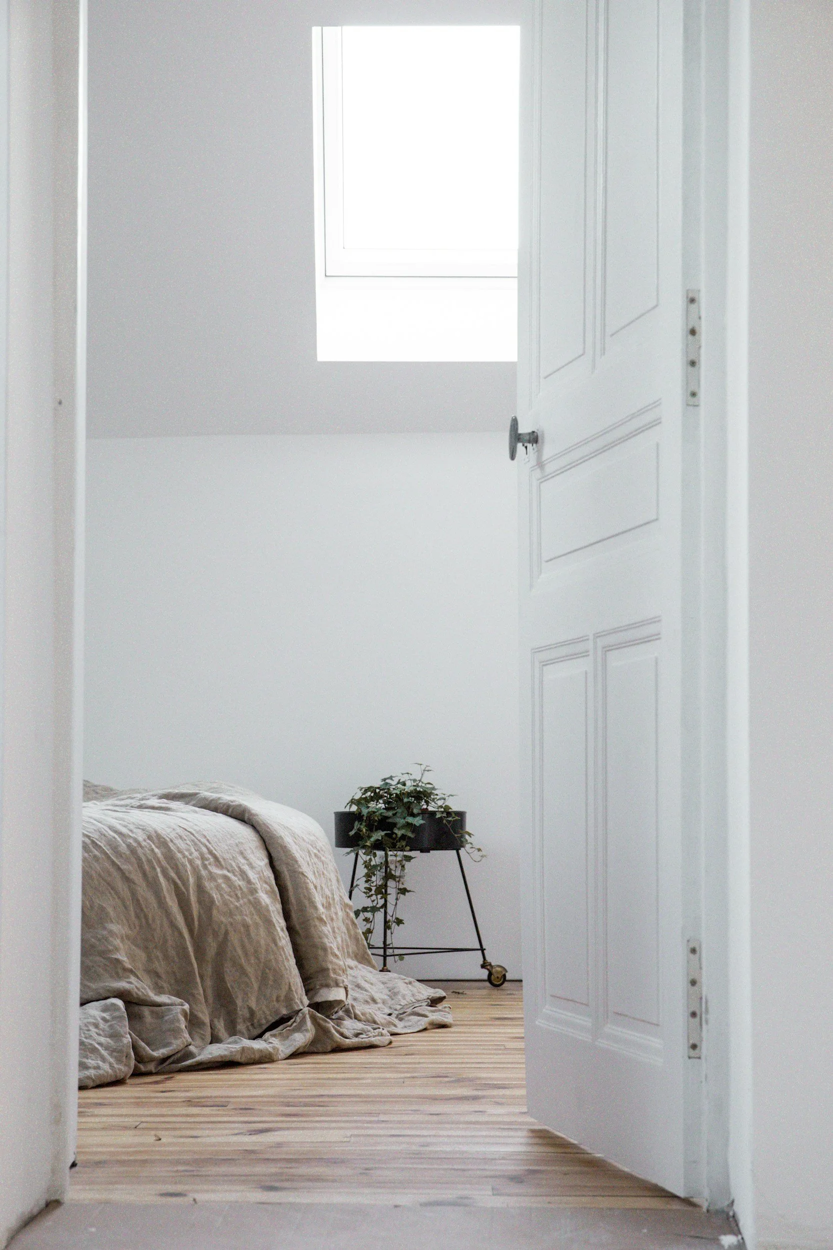 View of a bedroom through an open white door, with a bed covered in beige linen, a potted plant on a black stand, and a window on the back wall.
