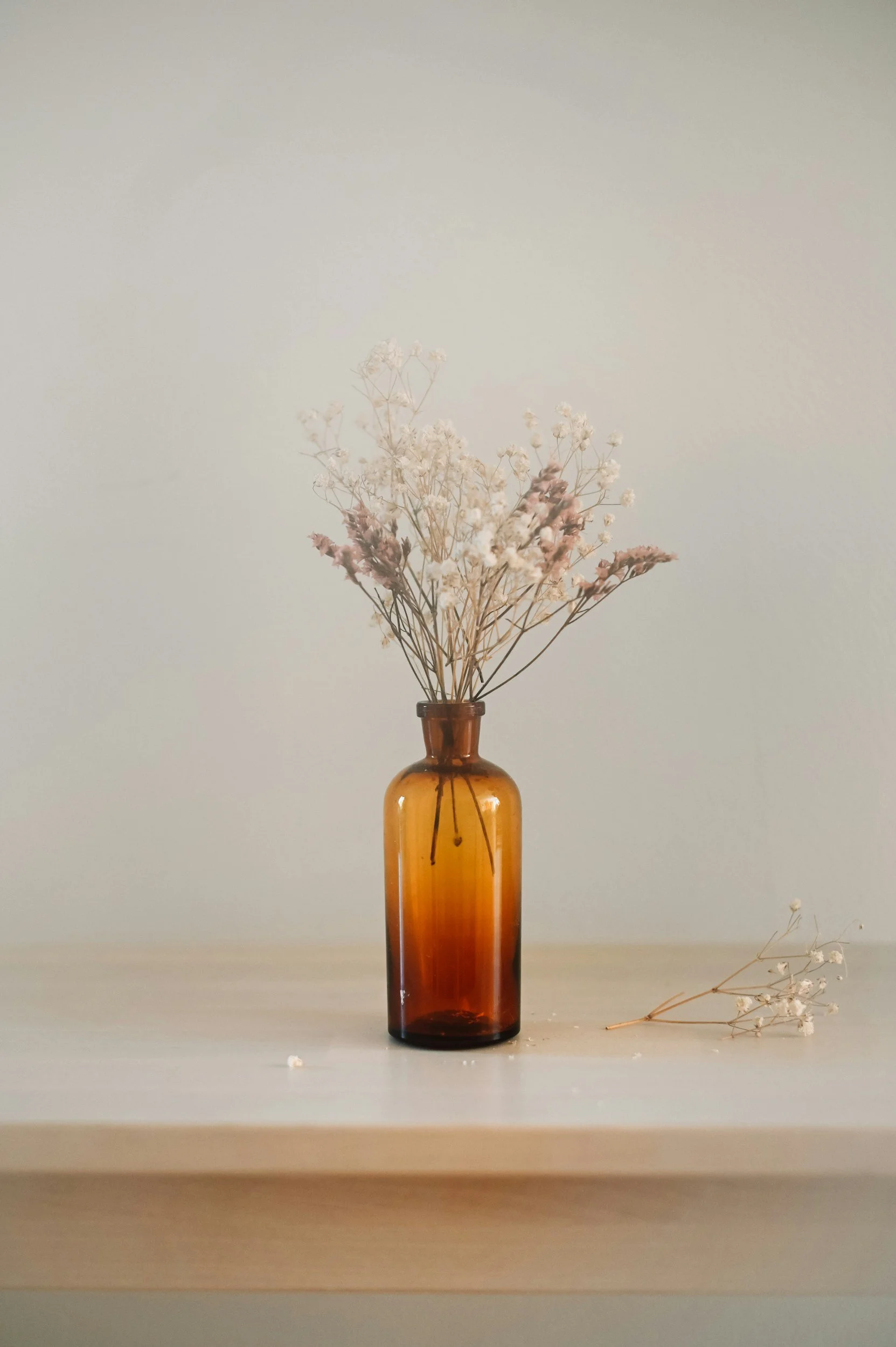 A brown glass vase with dried white and pink flowers on a light-colored surface against a plain background.
