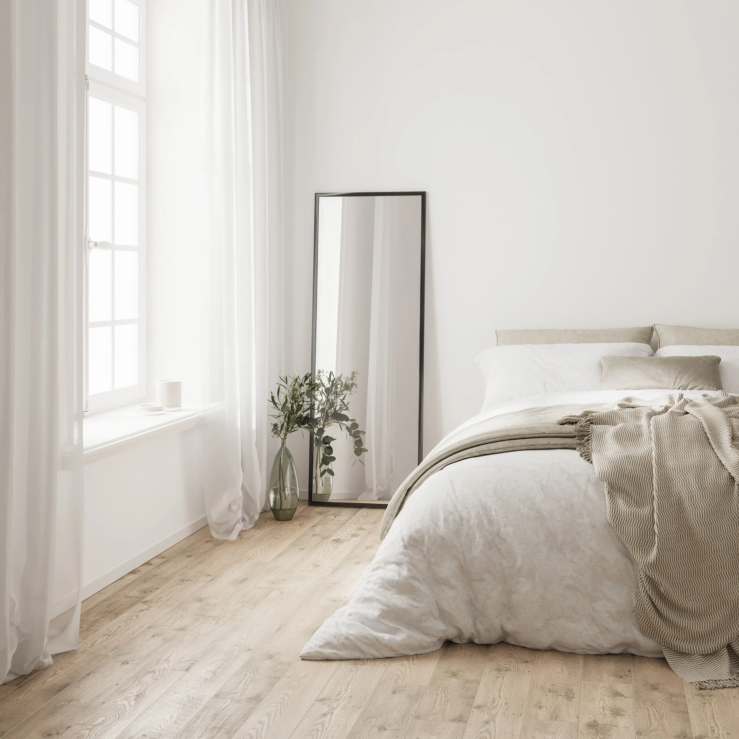 Bright bedroom with white walls, large window with sheer curtains, leaning full-length mirror, potted plant, unmade bed with beige and white bedding, and wooden flooring.
