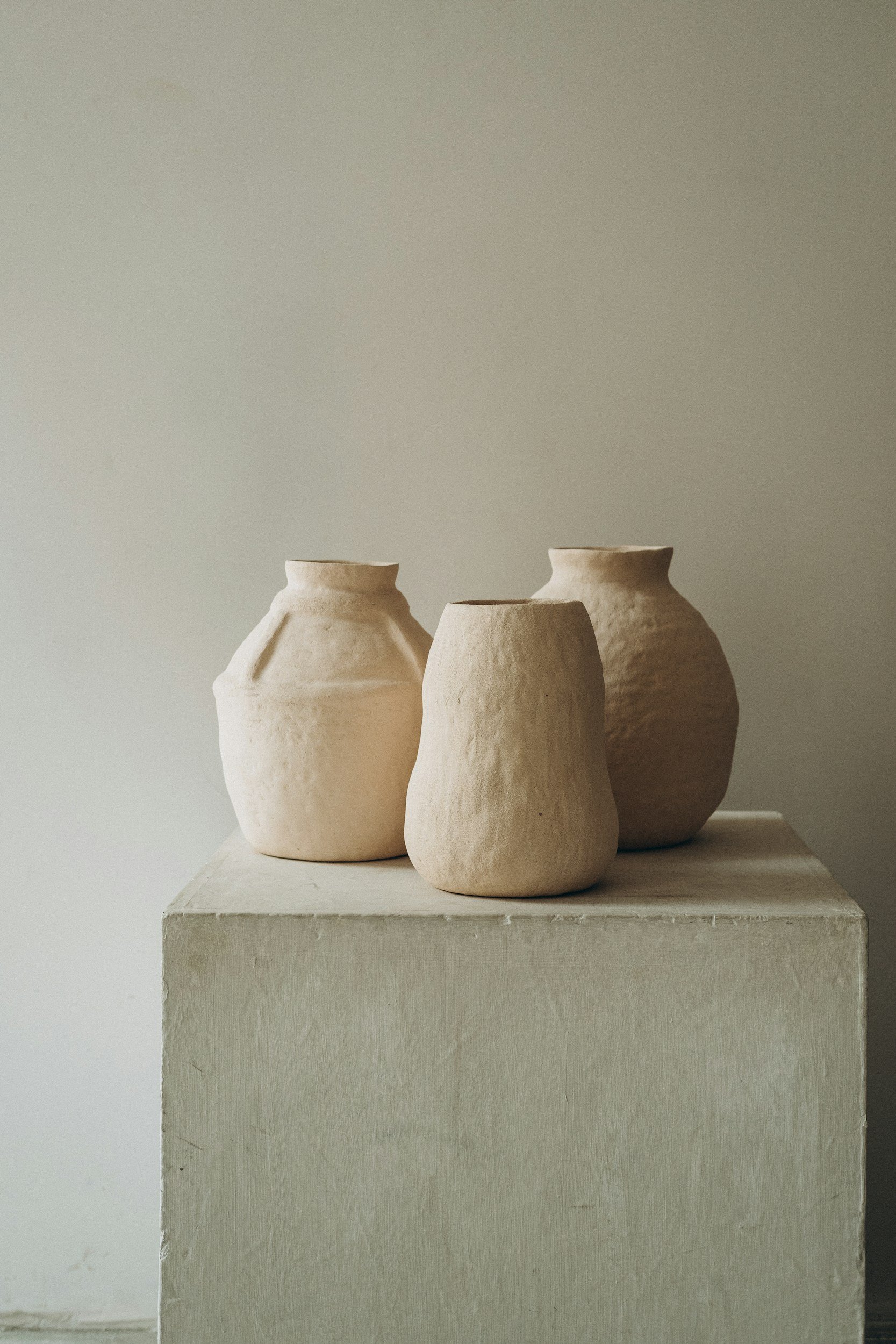 Three beige ceramic vases with textured surfaces on a white pedestal against a plain wall.