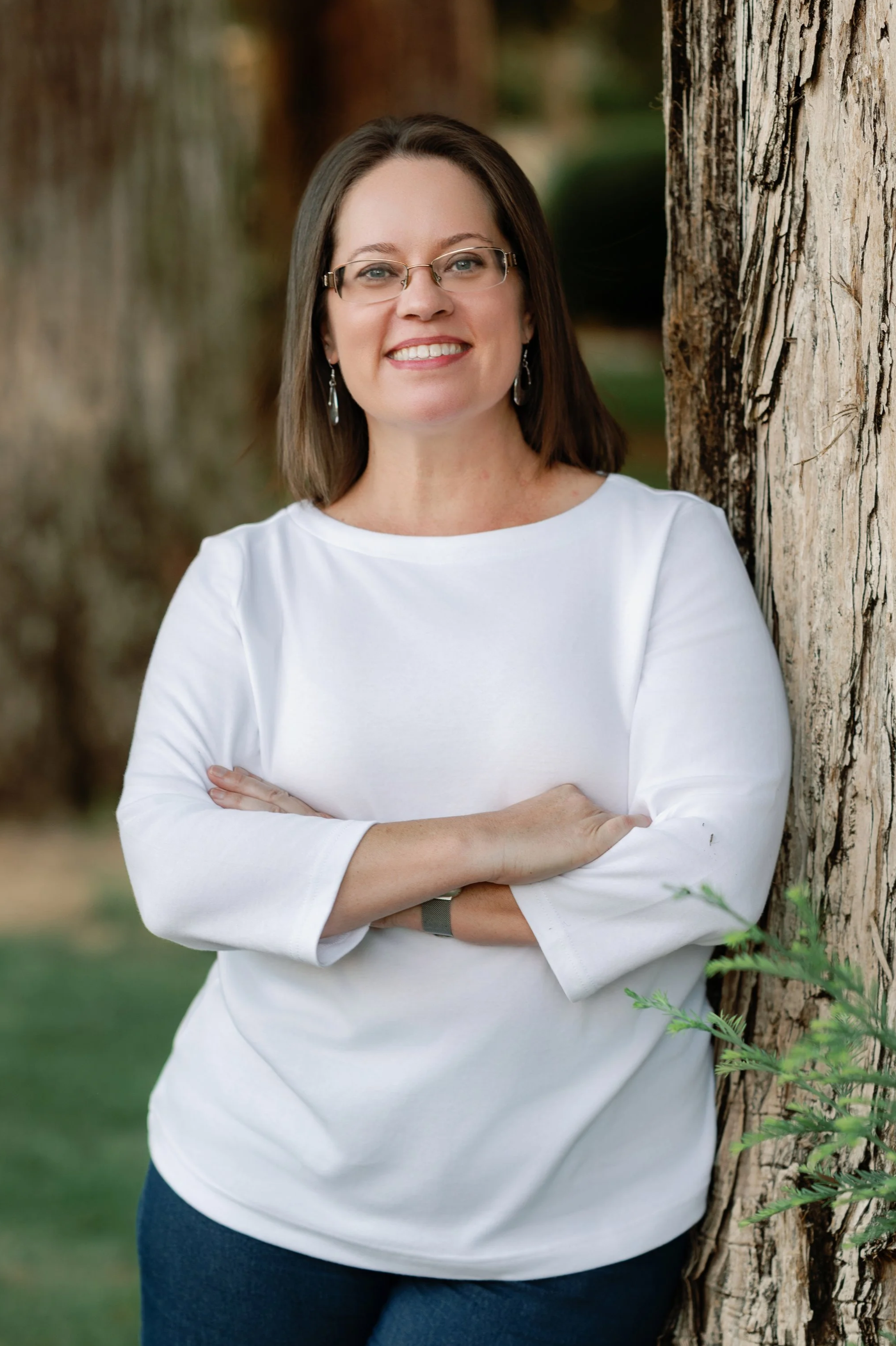 A woman with shoulder-length brown hair, glasses, and earrings is smiling and posing outdoors next to a tree, wearing a white top and dark pants.