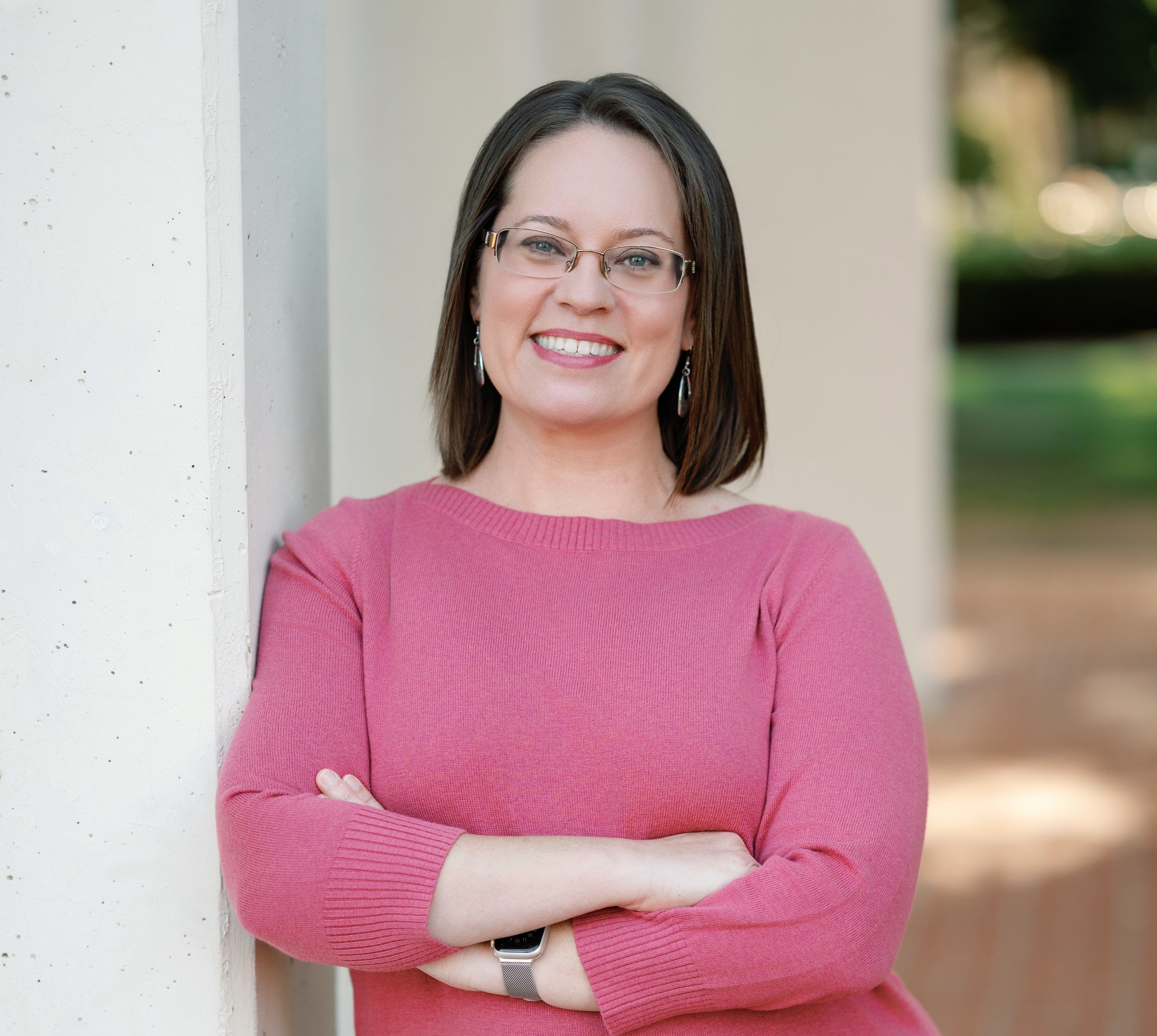 A woman with shoulder-length brown hair wearing a pink sweater, glasses, and earrings, smiling with arms crossed, standing outdoors with blurred greenery in the background.