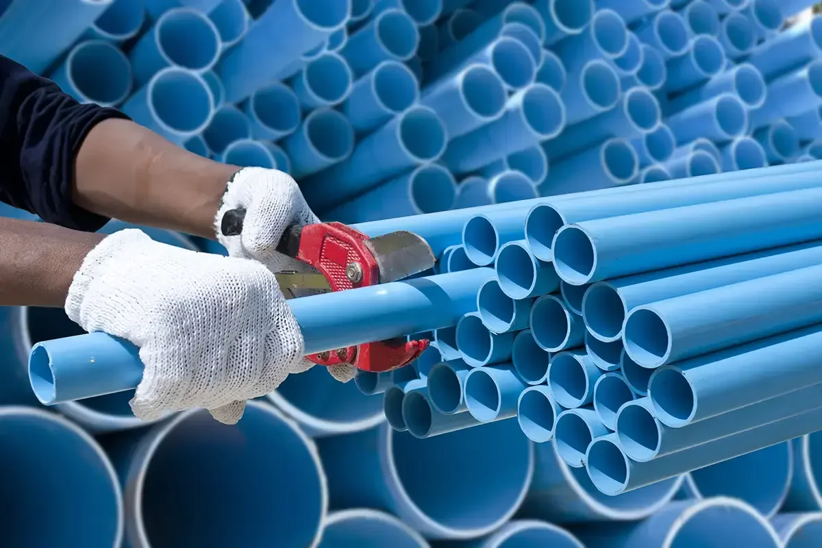 Person wearing white gloves cuts a blue plastic pipe with a cutter, with large stacks of similar pipes in the background.
