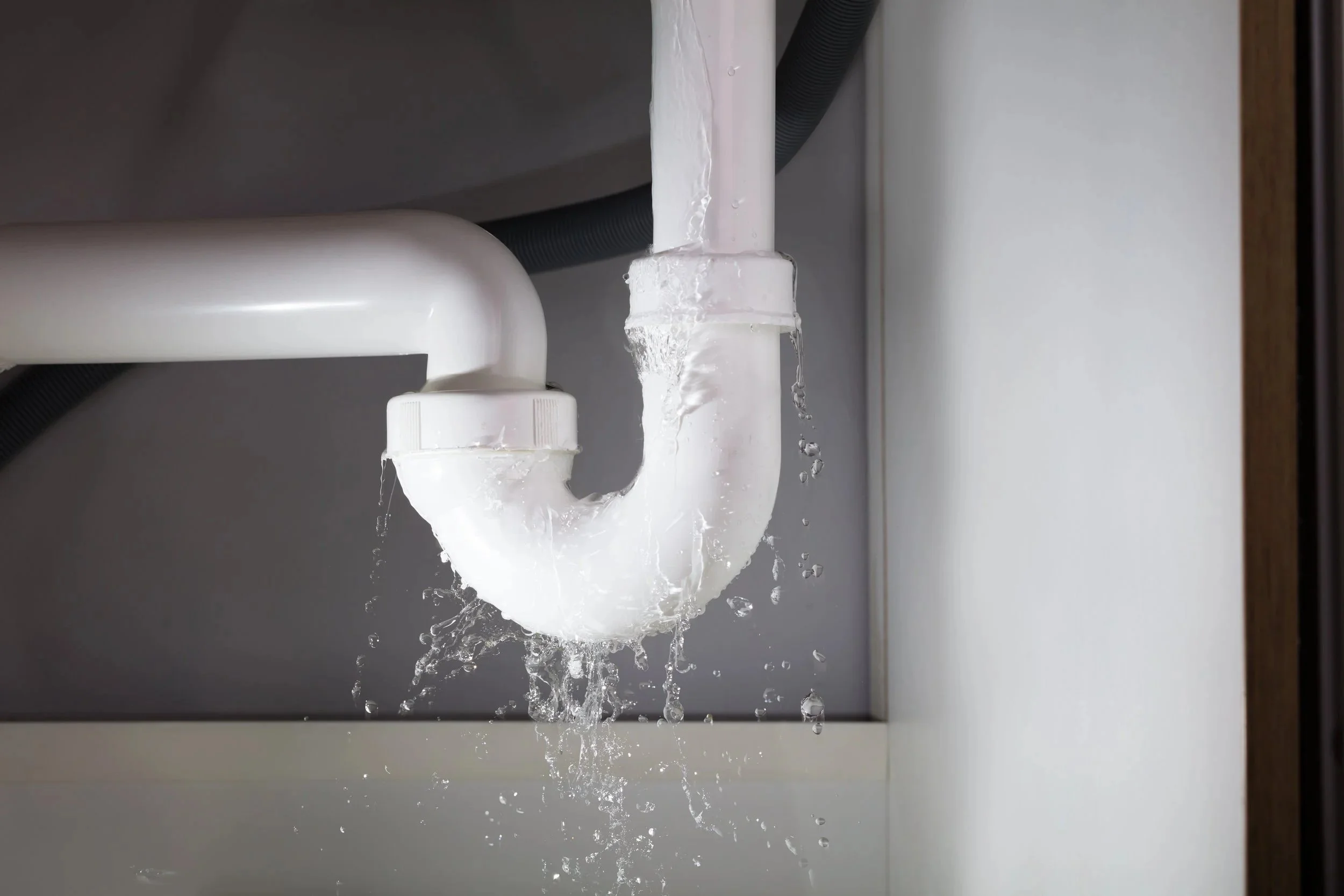 Water is flowing from a white PVC pipe under a sink, with some water splashing and dripping onto the surface below.