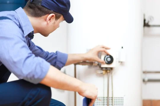 A man in a blue shirt and cap adjusting a boiler or water heater.