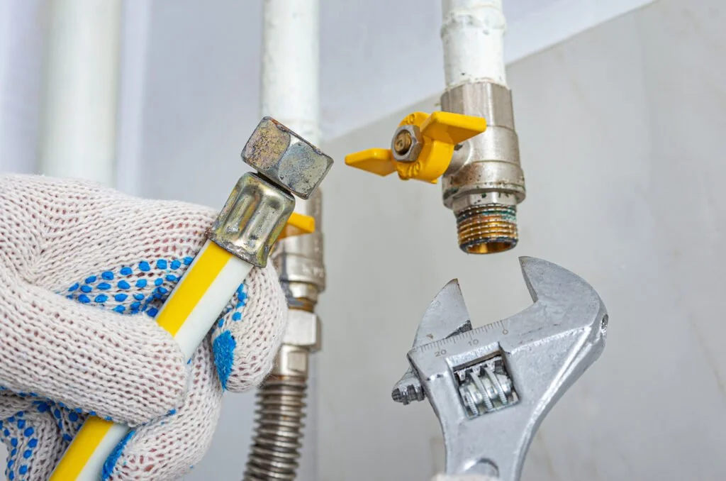 A person wearing a glove is holding a small pipe wrench near a plumbing faucet with a yellow handle. The faucet is attached to a wall and there is a larger adjustable wrench positioned below it, ready to turn.