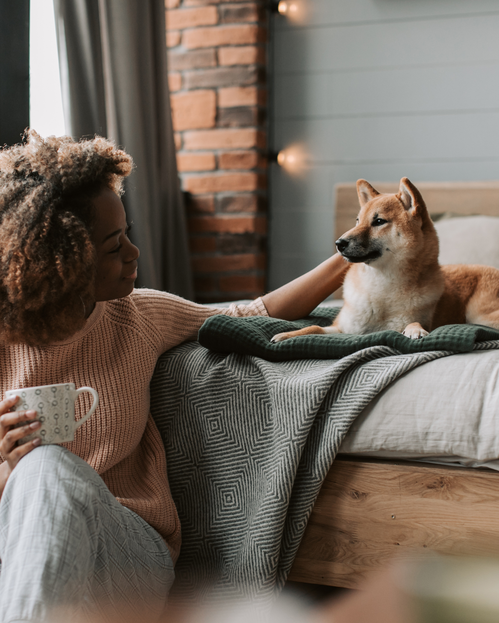 A woman with curly hair sitting on a bed holding a mug, gently petting a Shiba Inu dog lying on the bed, in a cozy bedroom with brick and wood wall accents.