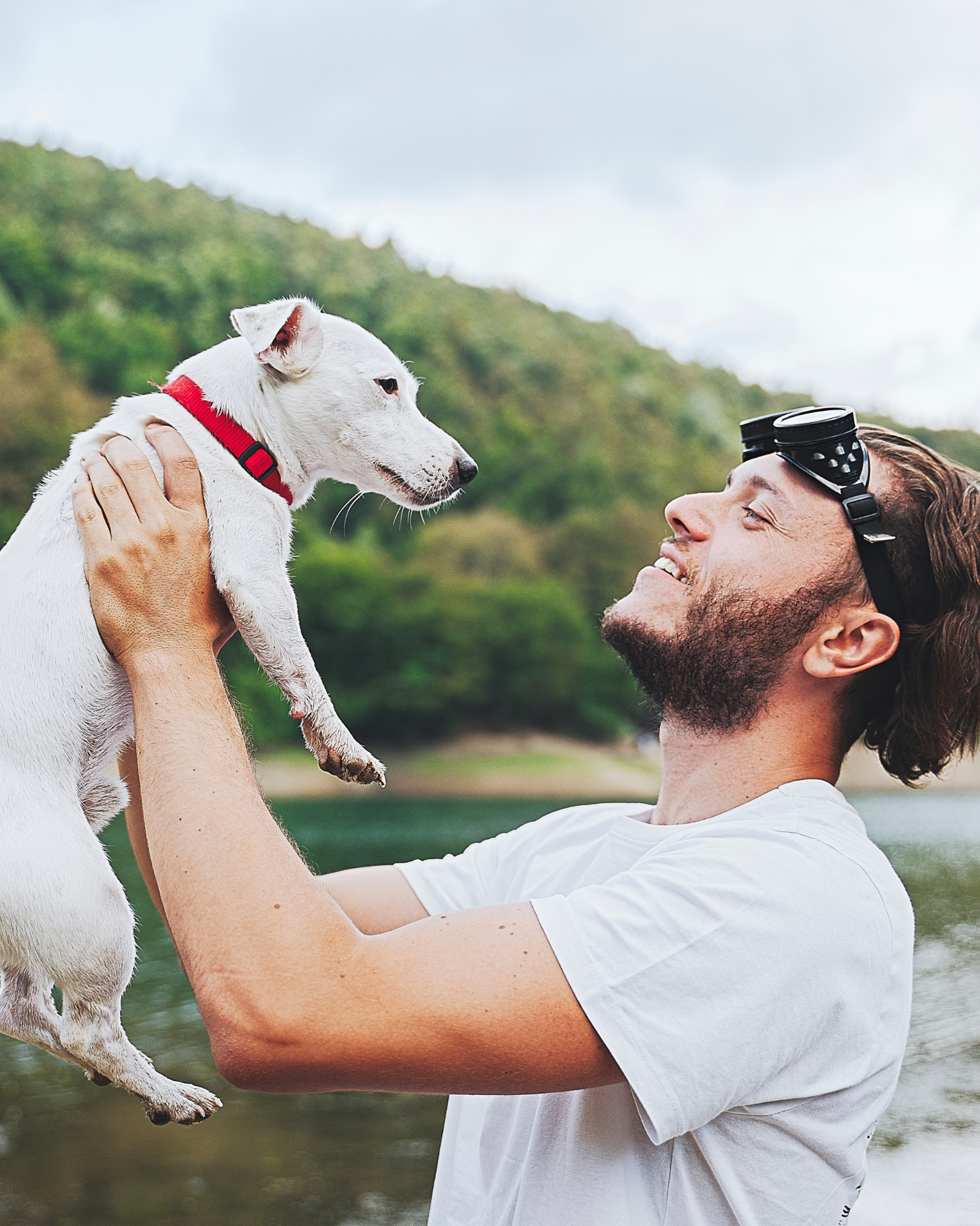 A man with a beard and sunglasses on his head is holding a small white dog with a red collar up in the air near a body of water, with green hills in the background.