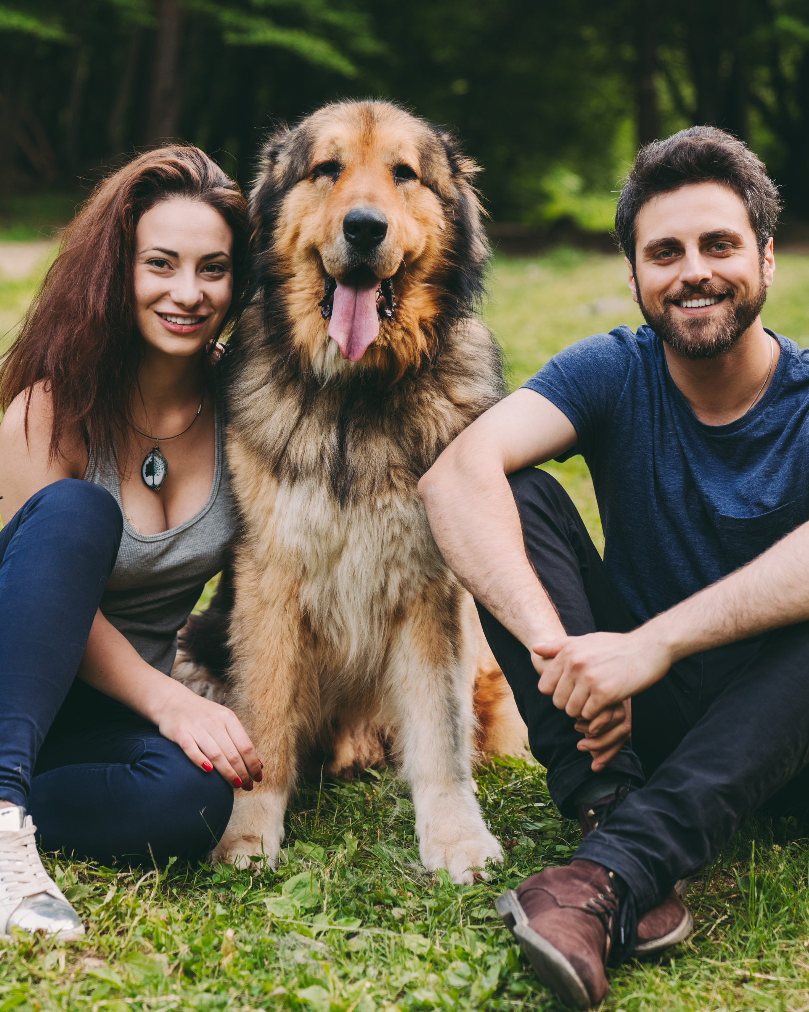 A woman and a man sitting on the grass next to a large, fluffy dog with a tan and black coat during daytime in a park.