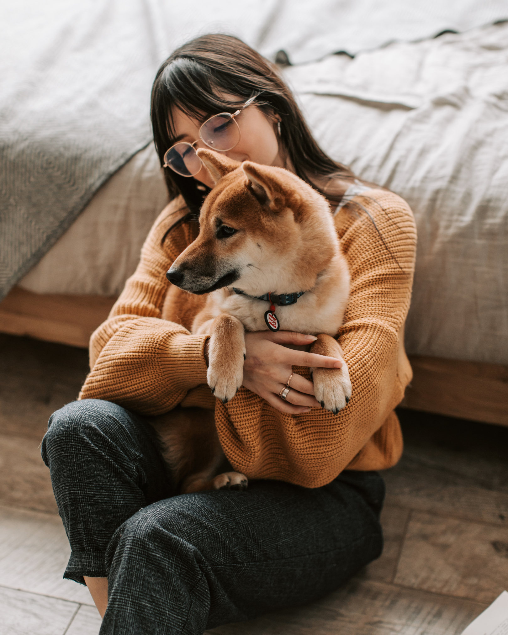 Young woman with glasses holding a Shiba Inu puppy while sitting on the floor near a bed.
