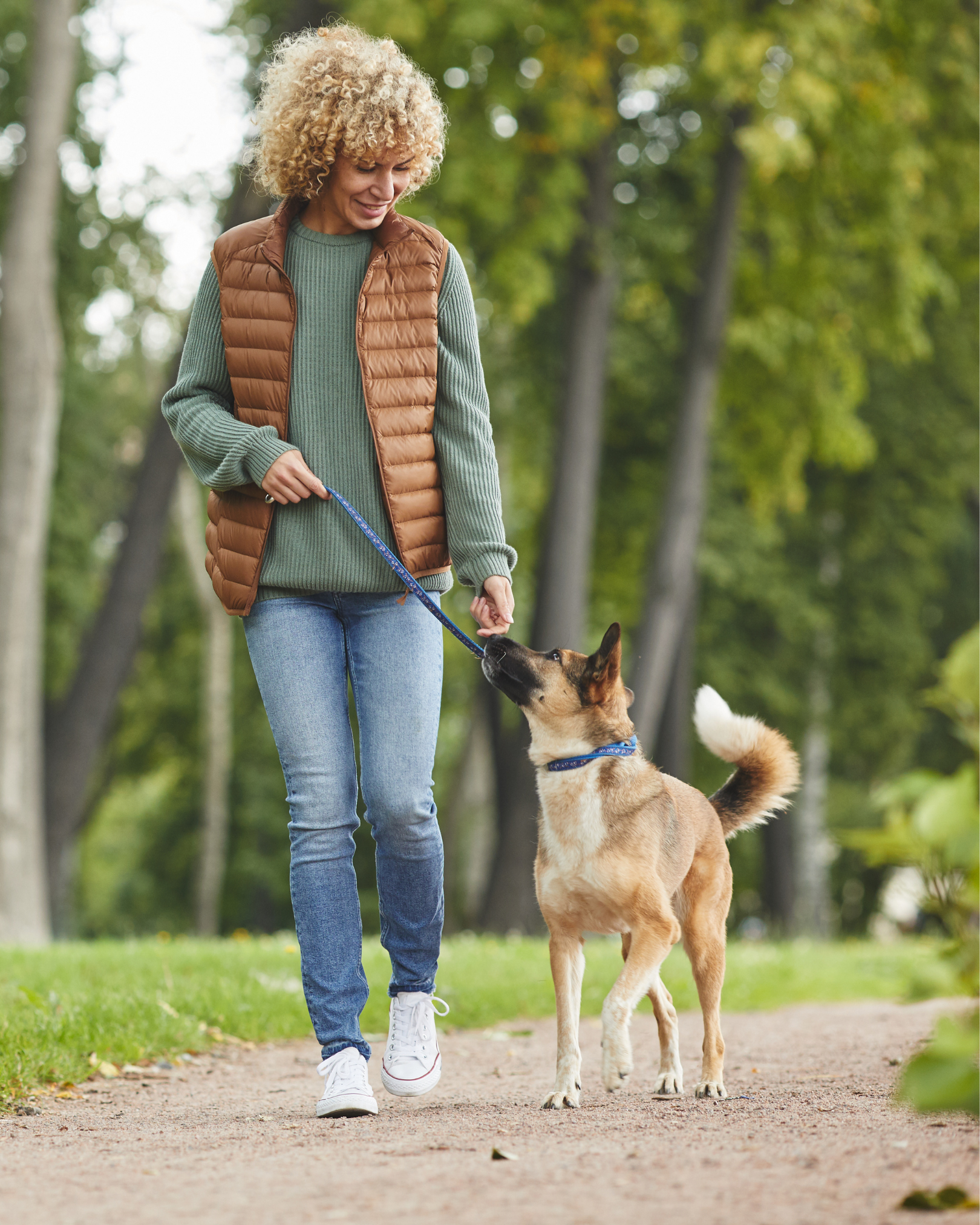 A woman with curly blonde hair wearing a green sweater, brown puffy vest, and blue jeans walking a dog on a leash in a park with green trees.
