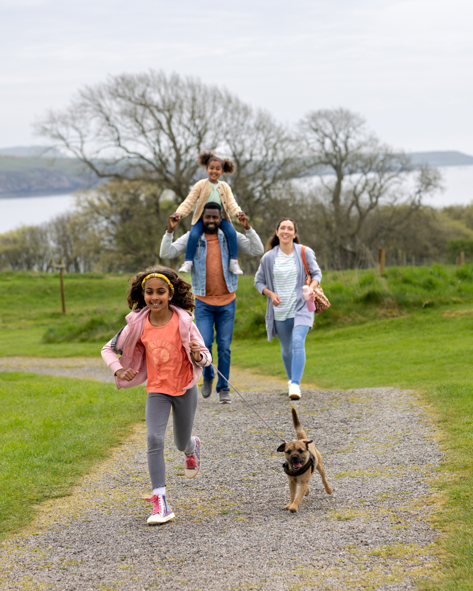 Group of people walking on a gravel path outdoors with a girl and dog in front, a man carrying a girl on his shoulders, and two women behind, in a green park with trees and lake in the background.