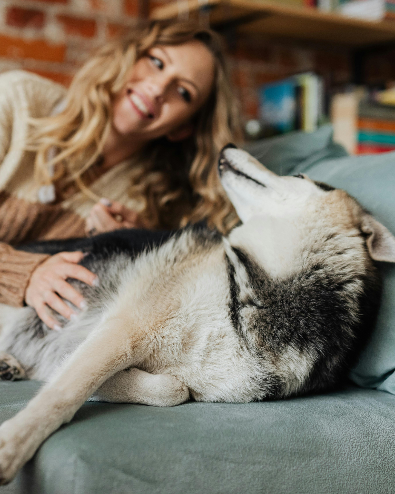 A woman with long, wavy blonde hair smiling while lying on a couch with a Siberian Husky dog, which is resting on its back with its nose touching her face.