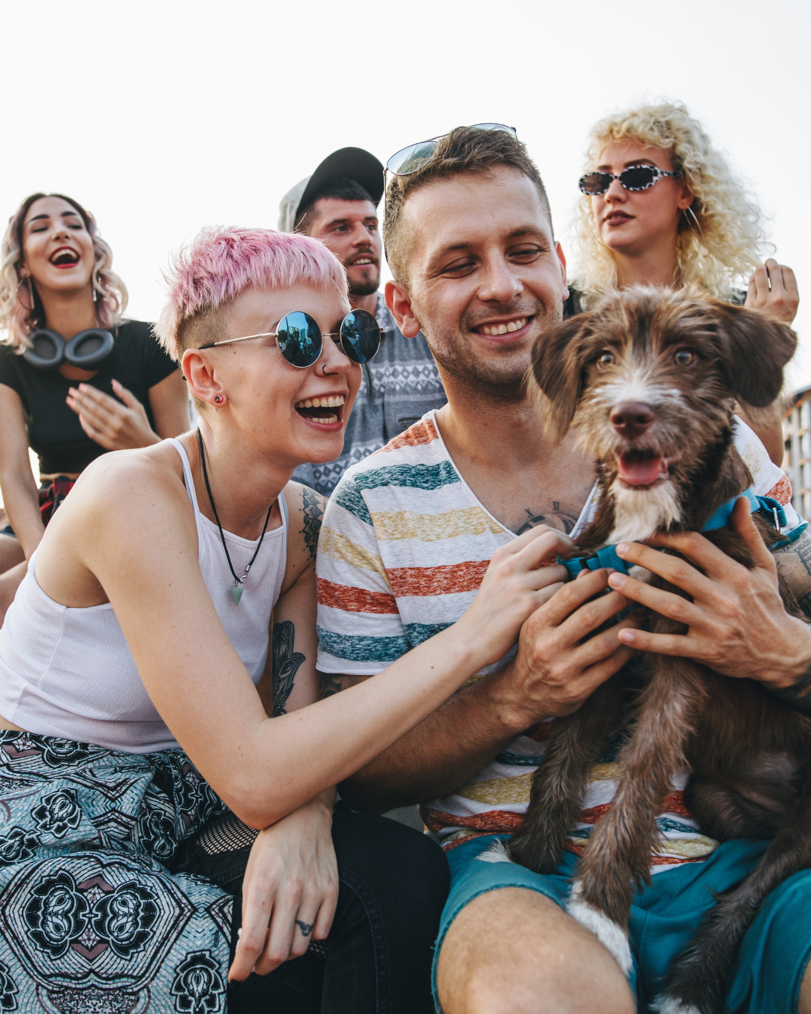 Group of friends smiling and laughing, holding a dog during an outdoor gathering.