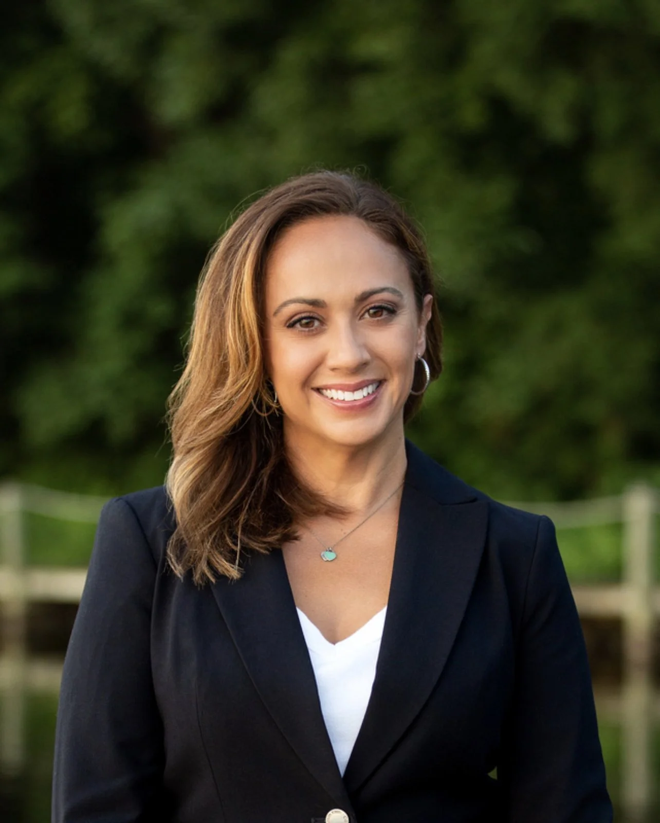 A woman with shoulder-length brown hair, smiling, wearing a black blazer, white top, earrings, and a necklace, standing outdoors with a blurred green background.