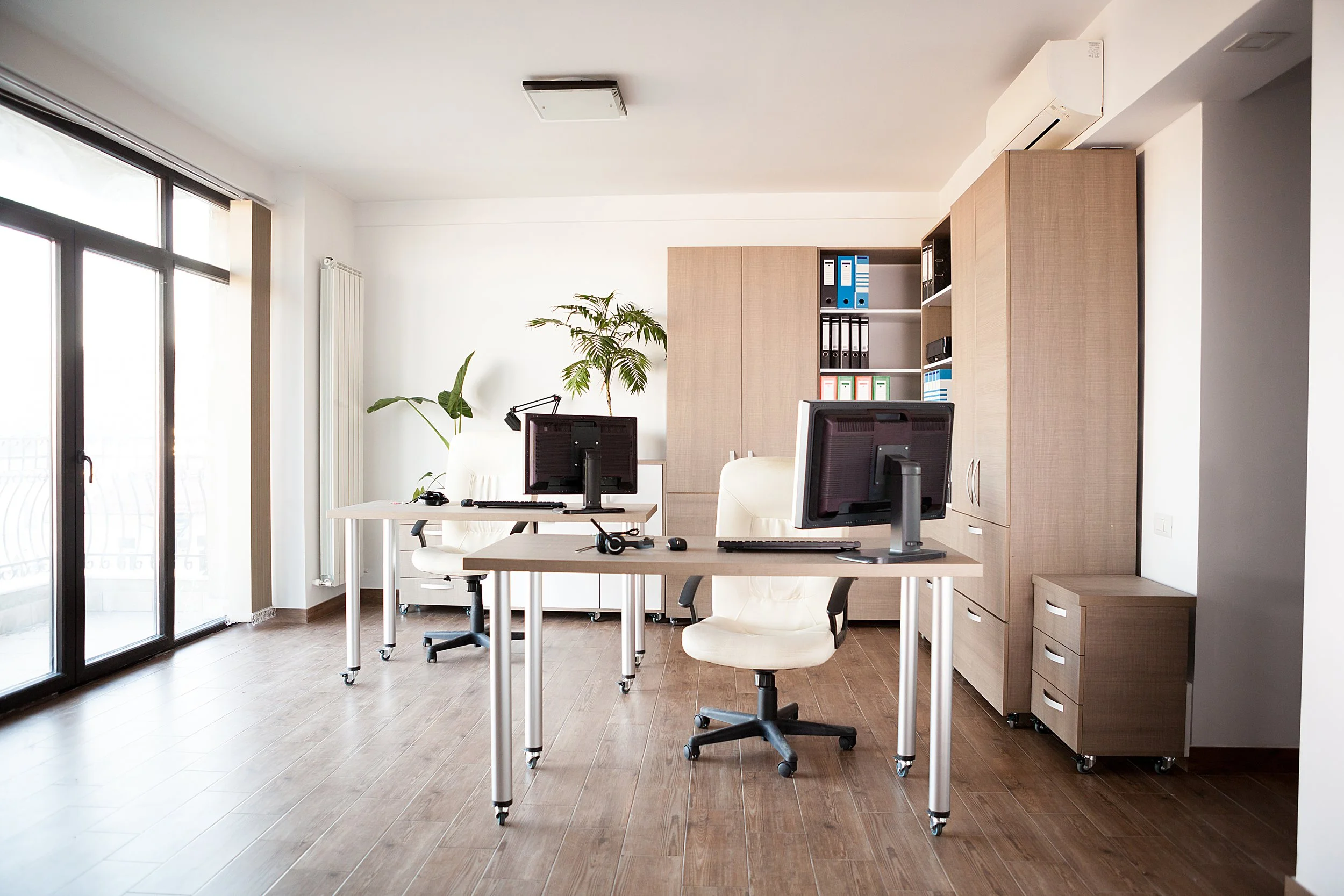 Modern office space with desks, computers, office chair, large window with sliding glass door, wooden flooring, and light-colored furniture.