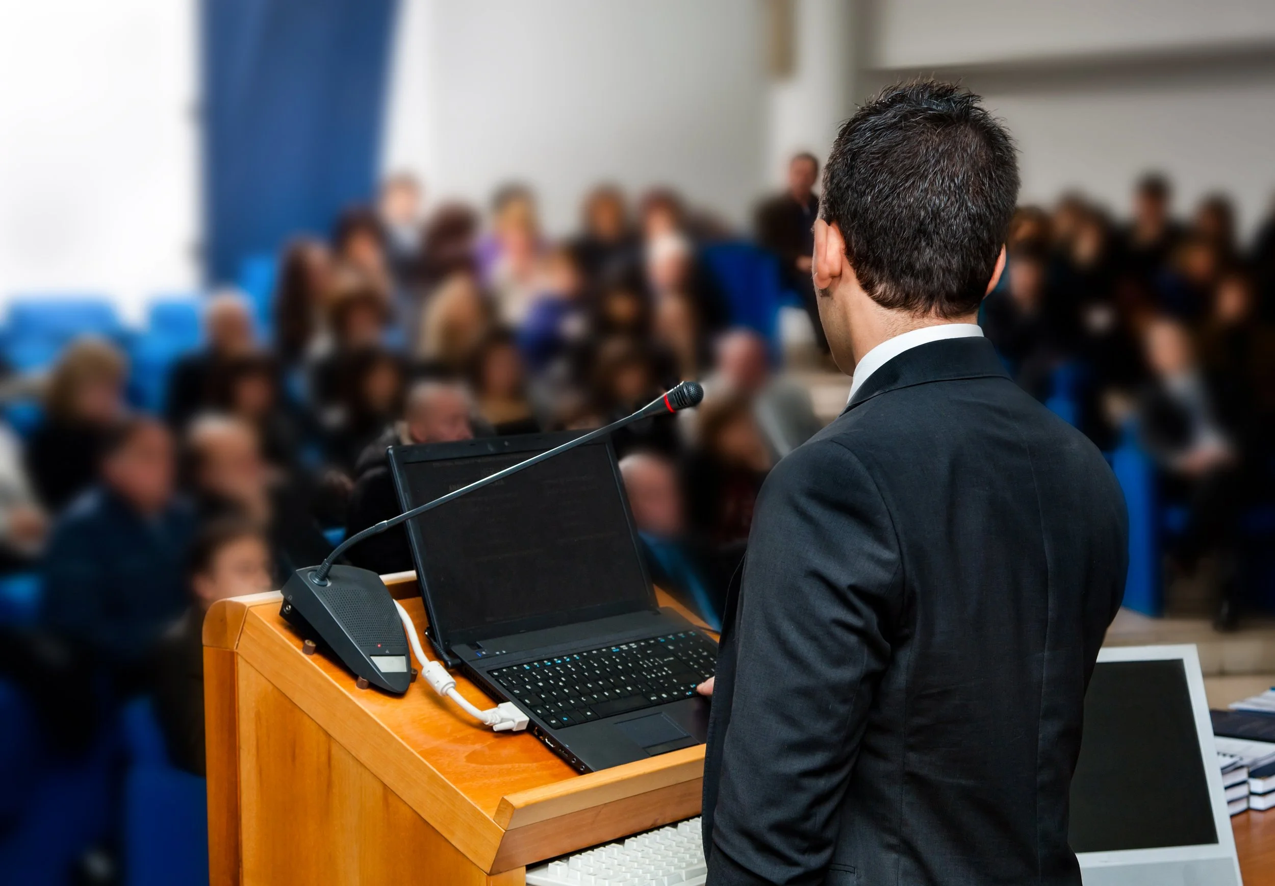 A man in a suit stands at a podium with a microphone, addressing an audience in a conference room.