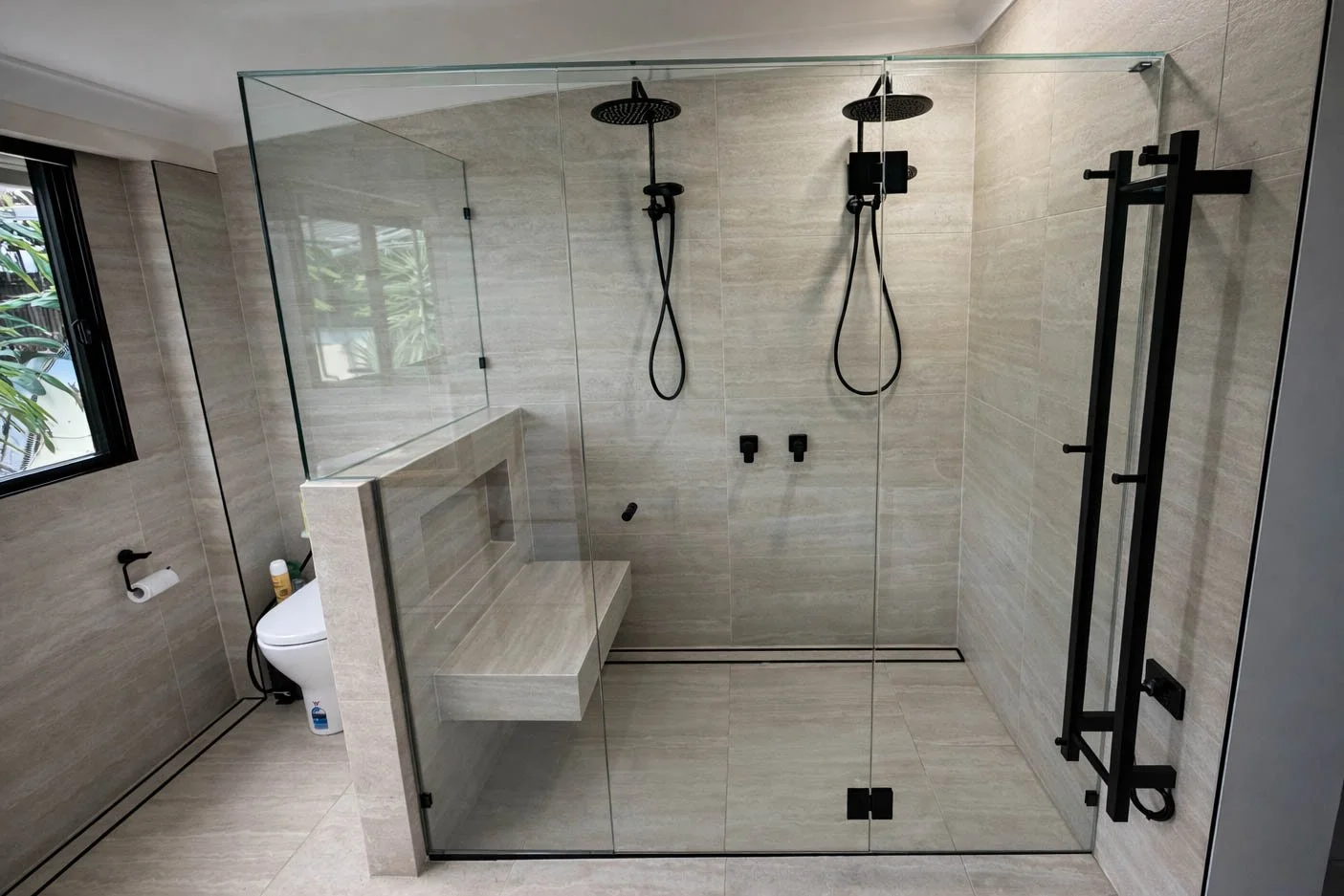 Modern shower area with two black showerheads and handhelds, glass enclosure, built-in bench, black fixtures, beige tiled walls, window, and towel rack.