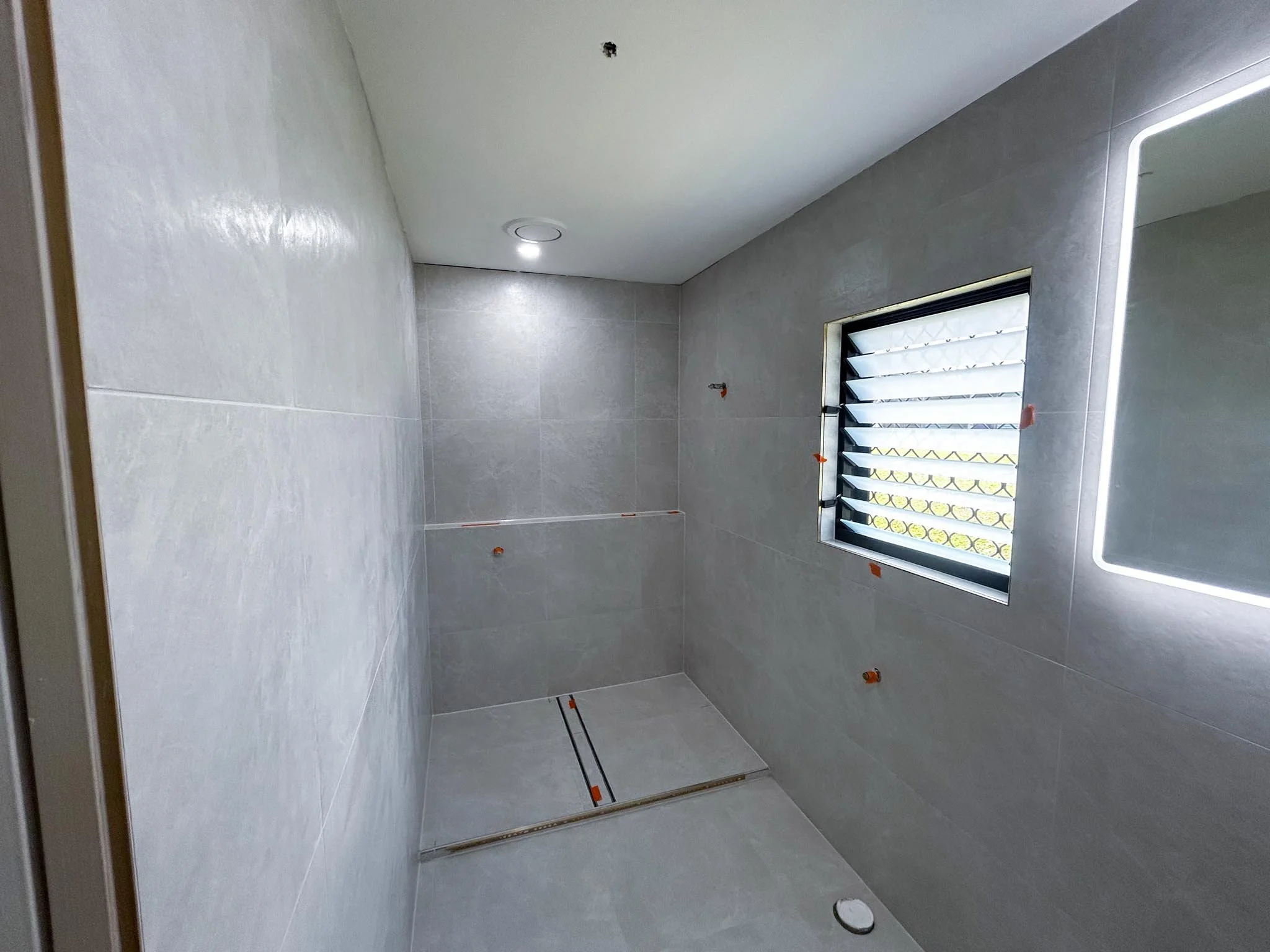 A modern bathroom under construction with grey tiled walls and a window with frosted glass. Small orange spacers are visible on the walls and floor for tile installation. There is a circular drainage cover on the floor, work by Birchwood Building Co.
