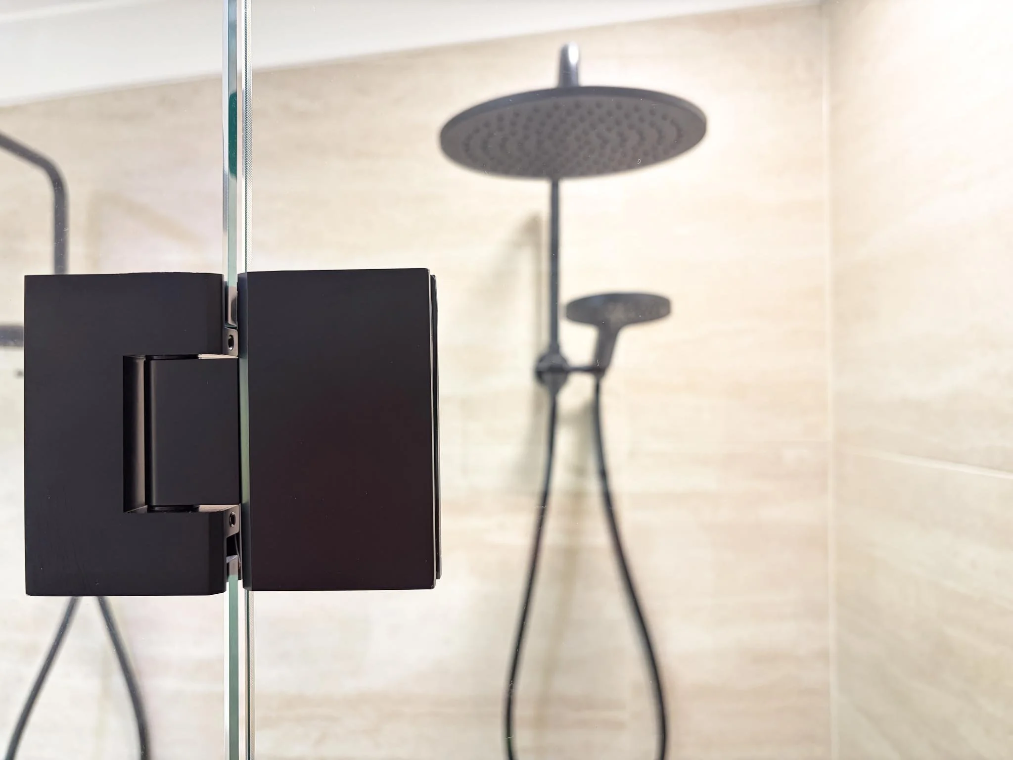 Close-up of a black glass shower door hinge with a modern shower head in the background showing a beige tiled shower interior.