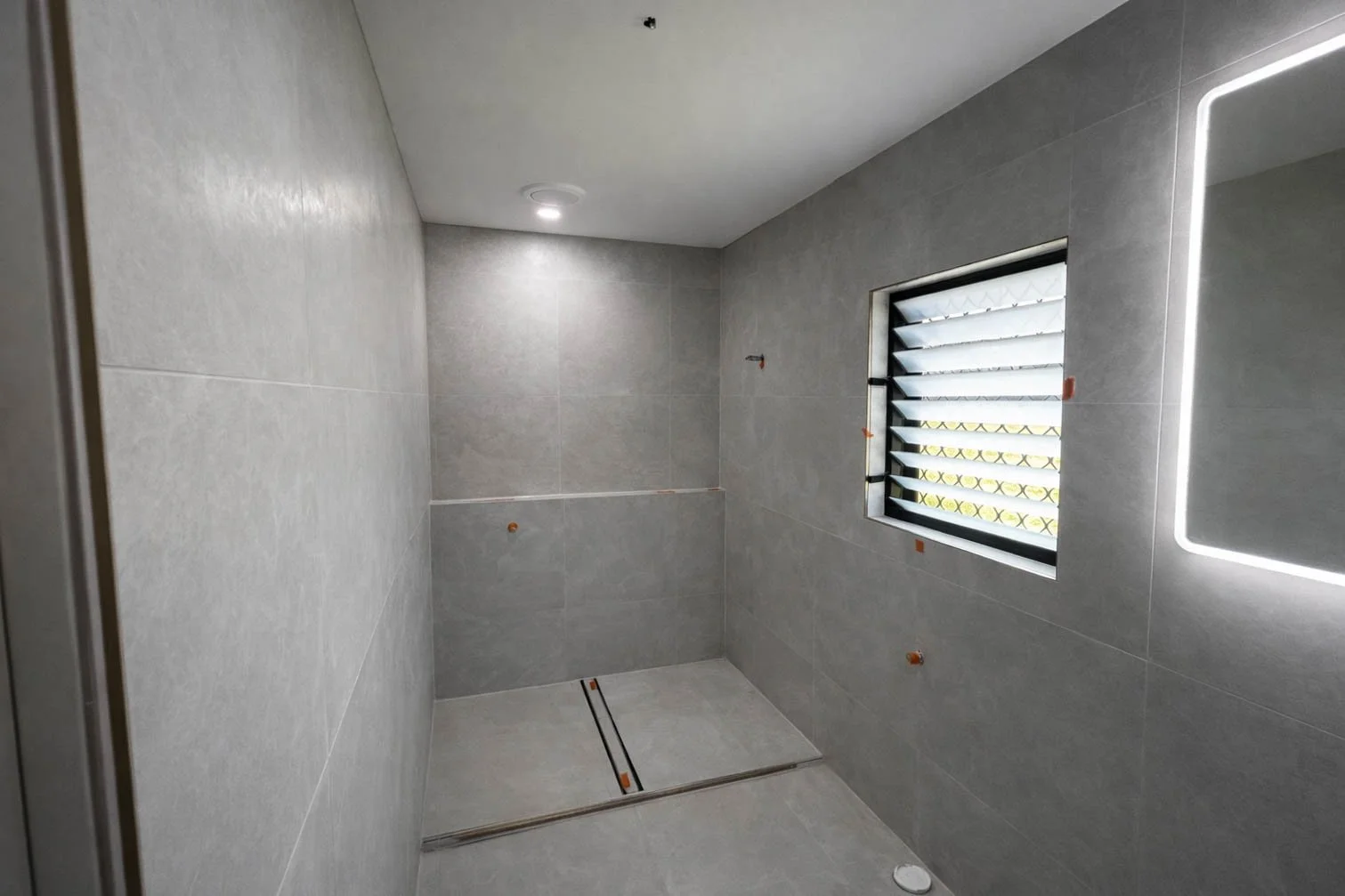 Empty modern shower with gray tiled walls and floor, a slatted window with black louvers, and a ceiling light, without fixtures or accessories.
