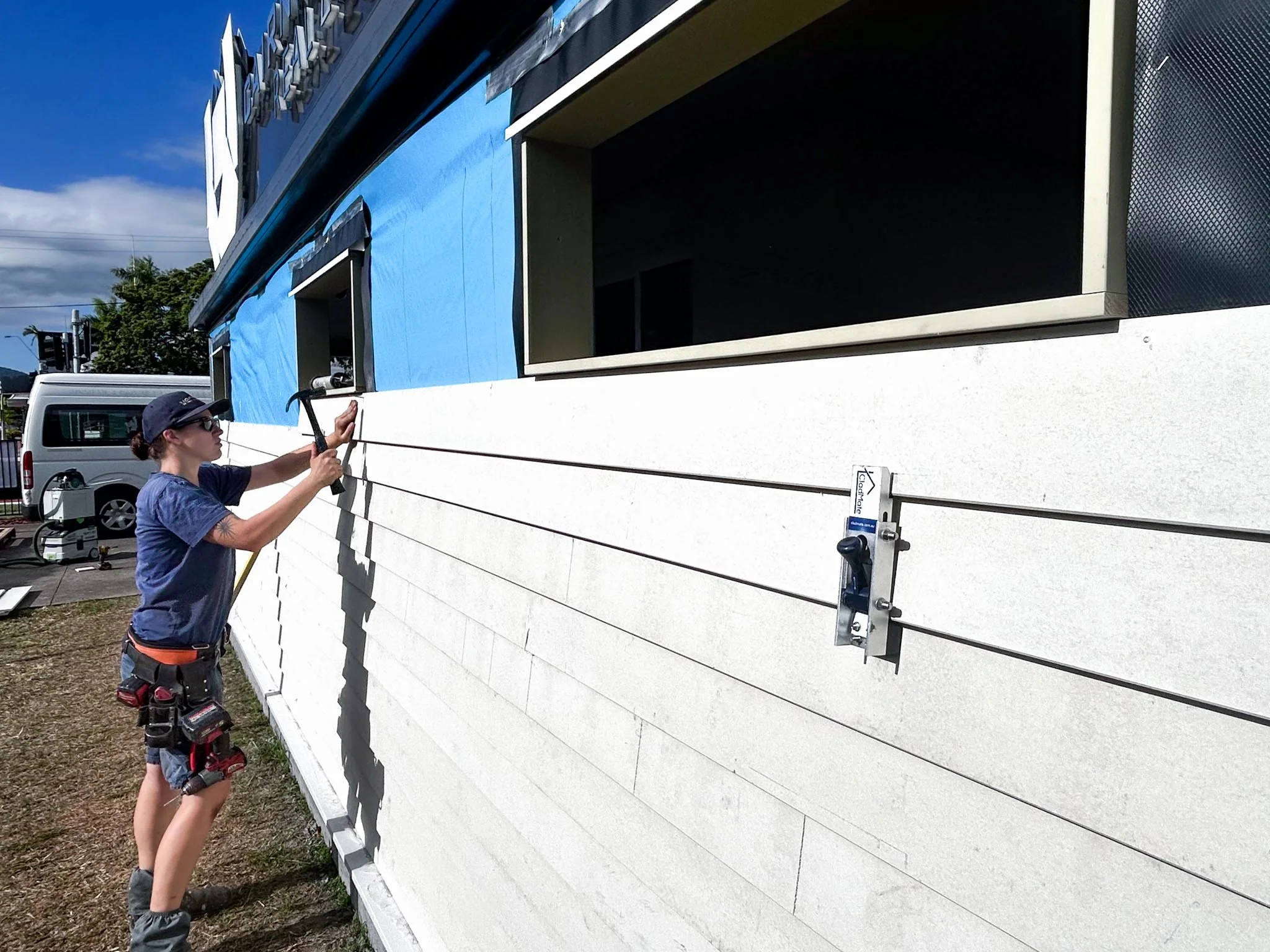 A woman working on the exterior of a building, using a hammer and a level, with tools attached to her belt. She is wearing a hat, sunglasses, a blue shirt, and shorts. The building has white siding with a window opening, and in the background, there is a white van and a blue sky.