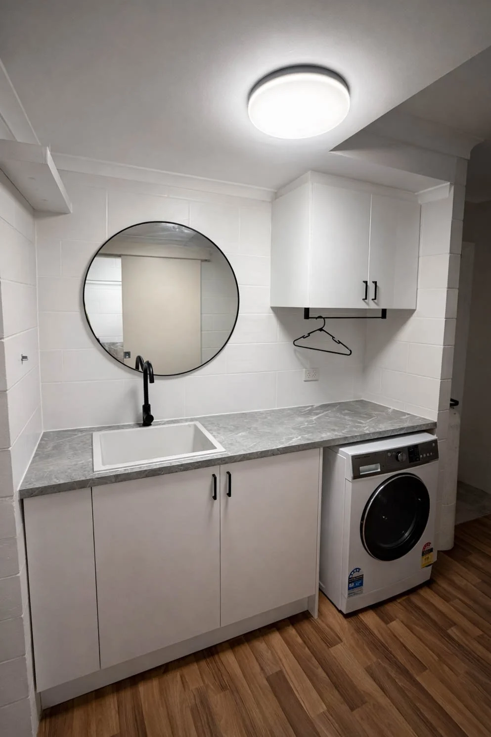 A laundry room with white cabinets, a marble countertop, a round mirror, a black faucet, a sink, a washing machine, and a cabinet with a hanger beneath it.