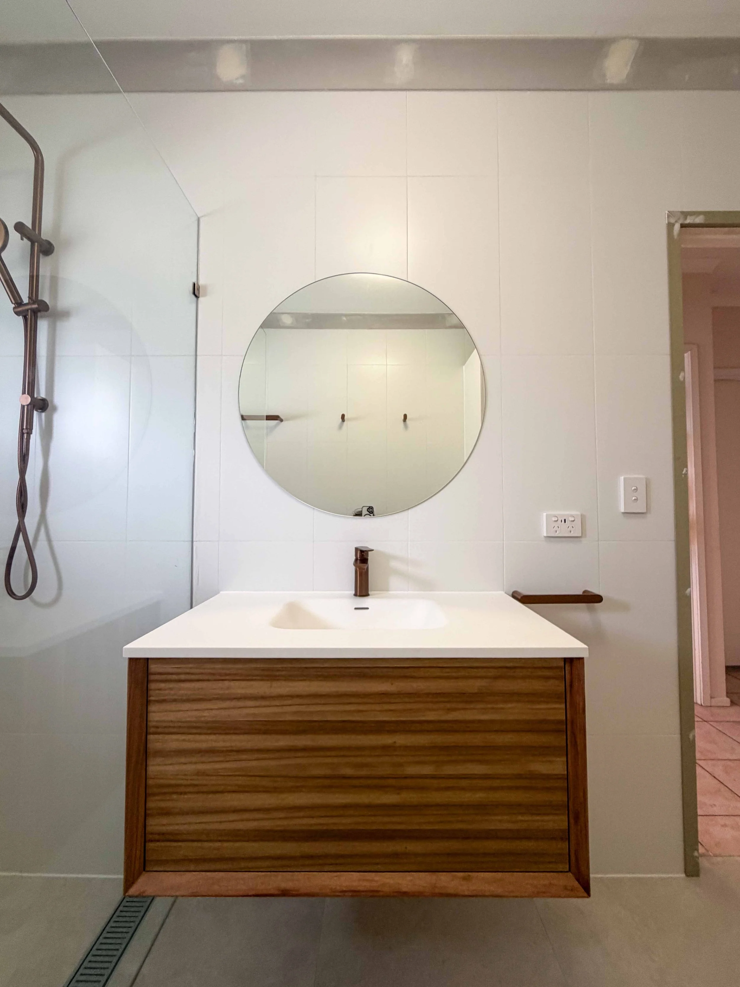 Bathroom with a wooden vanity, white countertop, round mirror, wall-mounted faucet, and a shower with a glass door and showerhead on the left.
