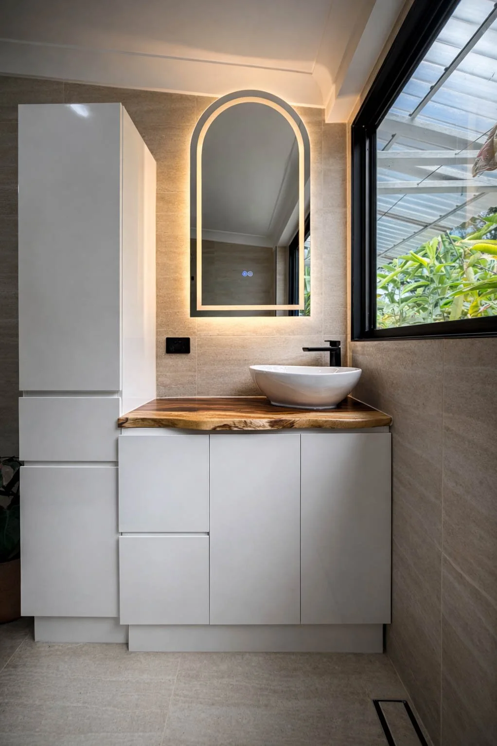 Modern bathroom vanity with white cabinets, a wood countertop, a vessel sink, a black faucet, a backlit mirror, beige tiled walls, and a large window showing outdoor greenery.