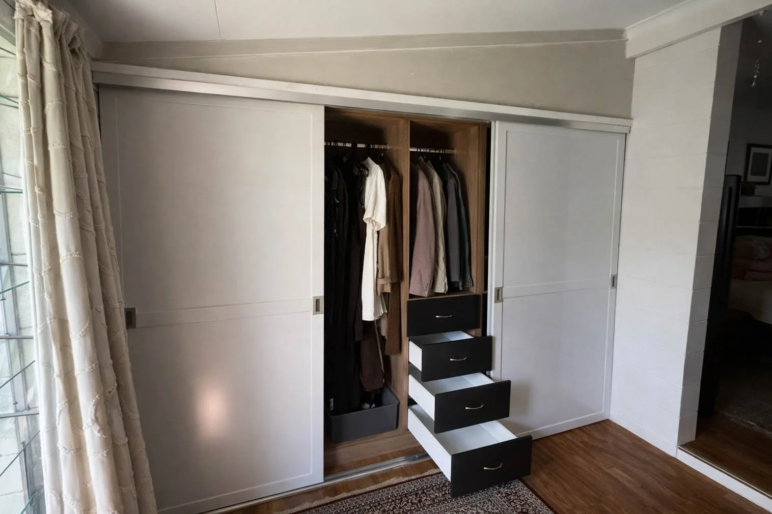 A built-in closet with sliding doors, partially open, revealing hanging clothes and drawers inside. The section with open drawers is black and white, with clothes hanging in the background. There is a window with beige curtains to the left and a hardwood floor with a rug in front.
