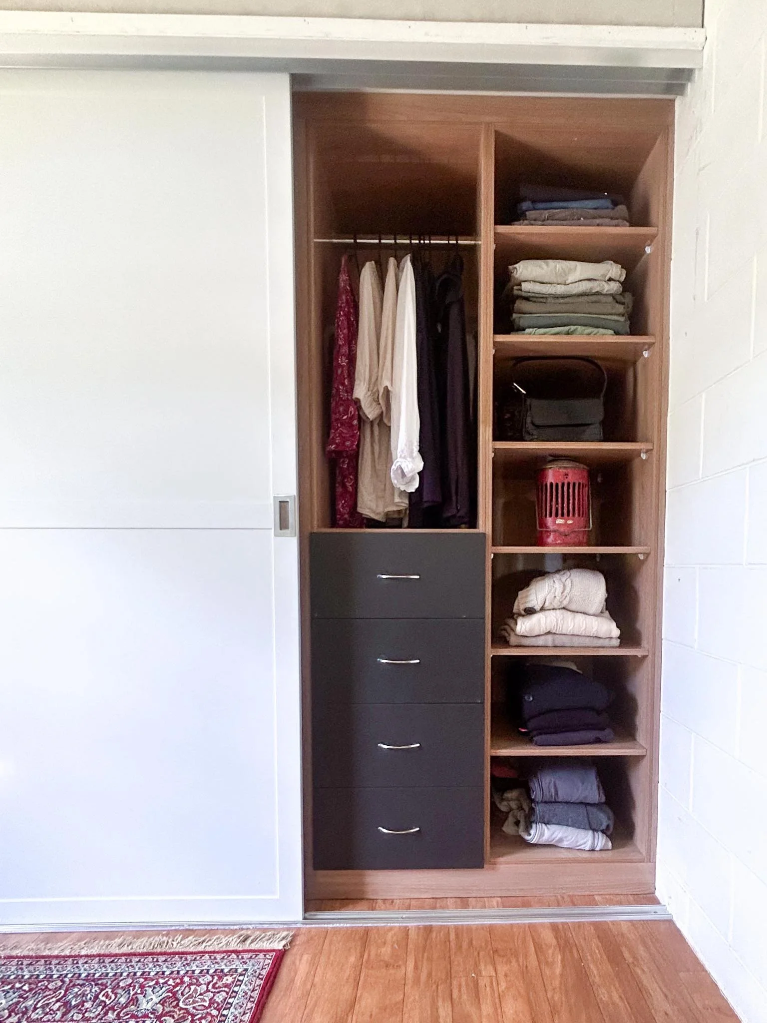 Closet with hanging clothes, folded clothes on shelves, a black bag, a red container, and drawers, with a wooden floor and a white sliding door.
